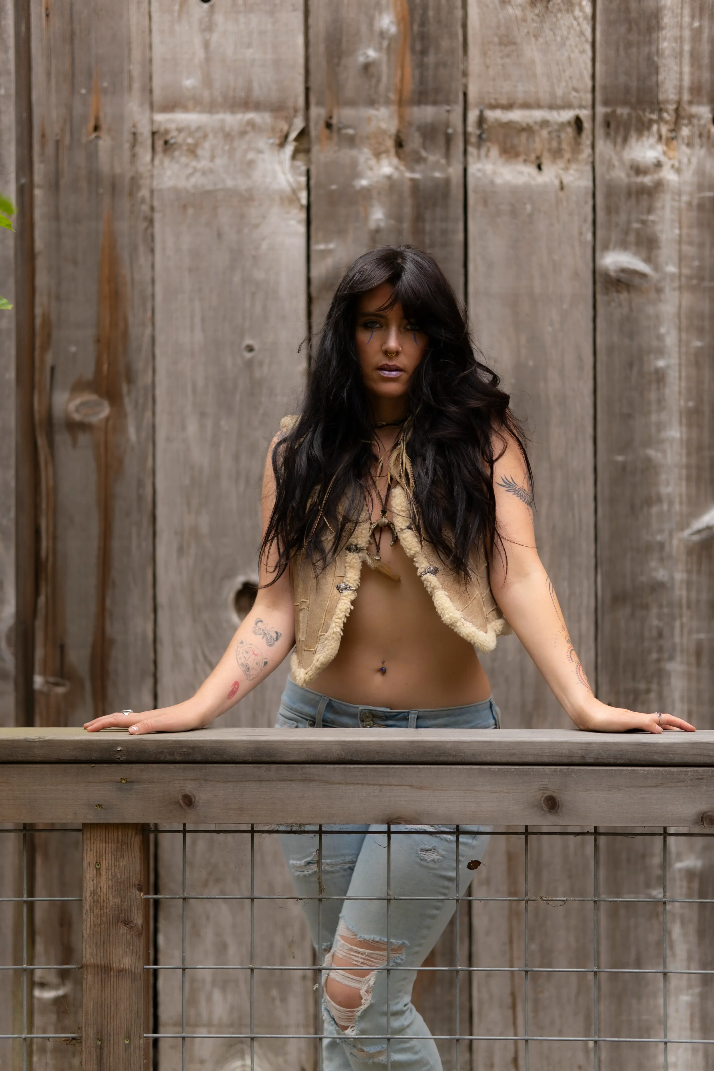 A young woman with long black hair and tattoos on her arms and stomach stands behind a wooden and metal railing against a wooden fence background. She is wearing a beige cropped vest and ripped jeans, with her hands resting on the railing.