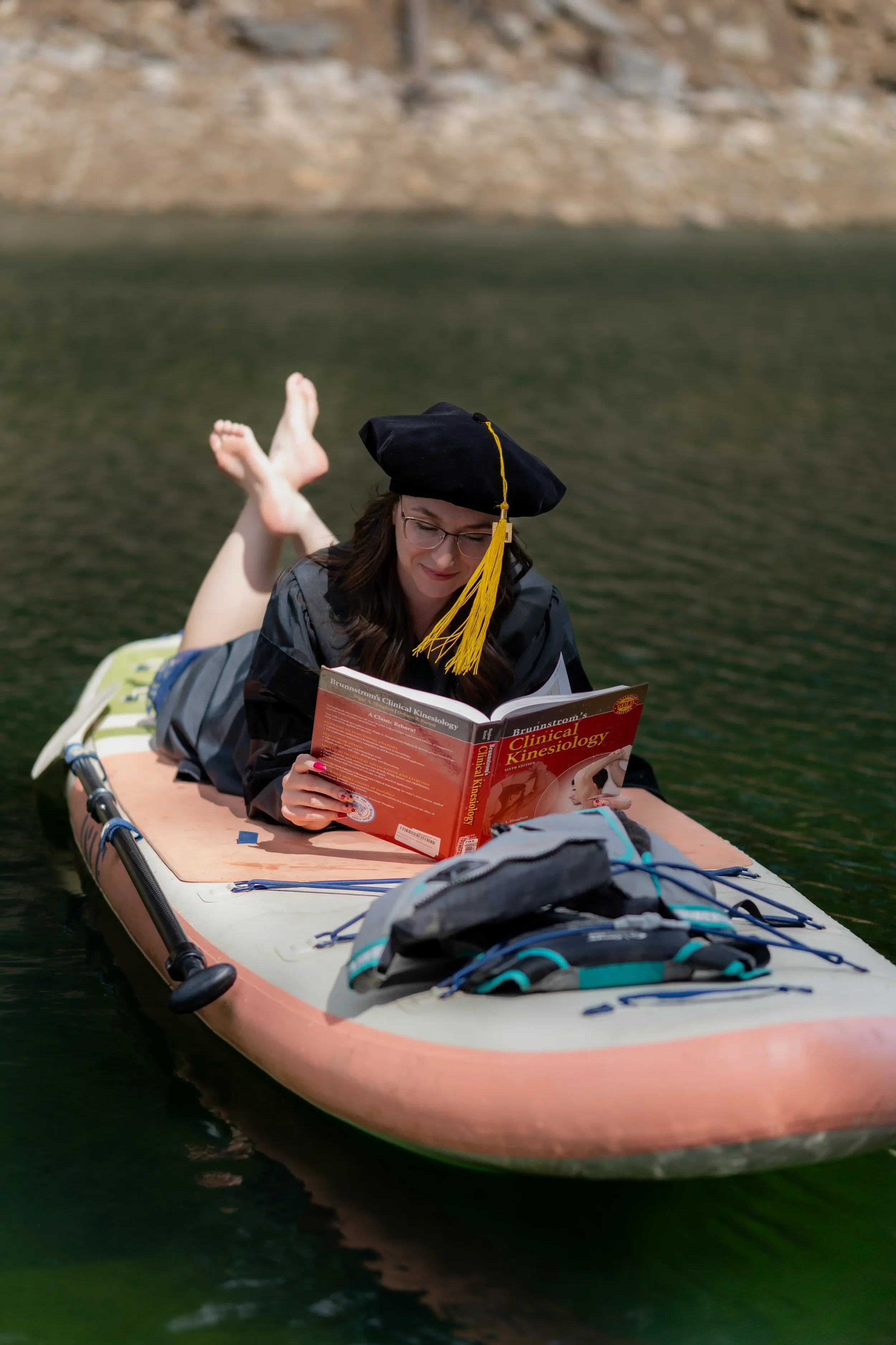A woman in graduation attire, including a black cap with a yellow tassel, is lying on her stomach on a paddleboard on a lake, reading a book titled 'Brunnstrom's Clinical Kinesiology' with trees in the background.