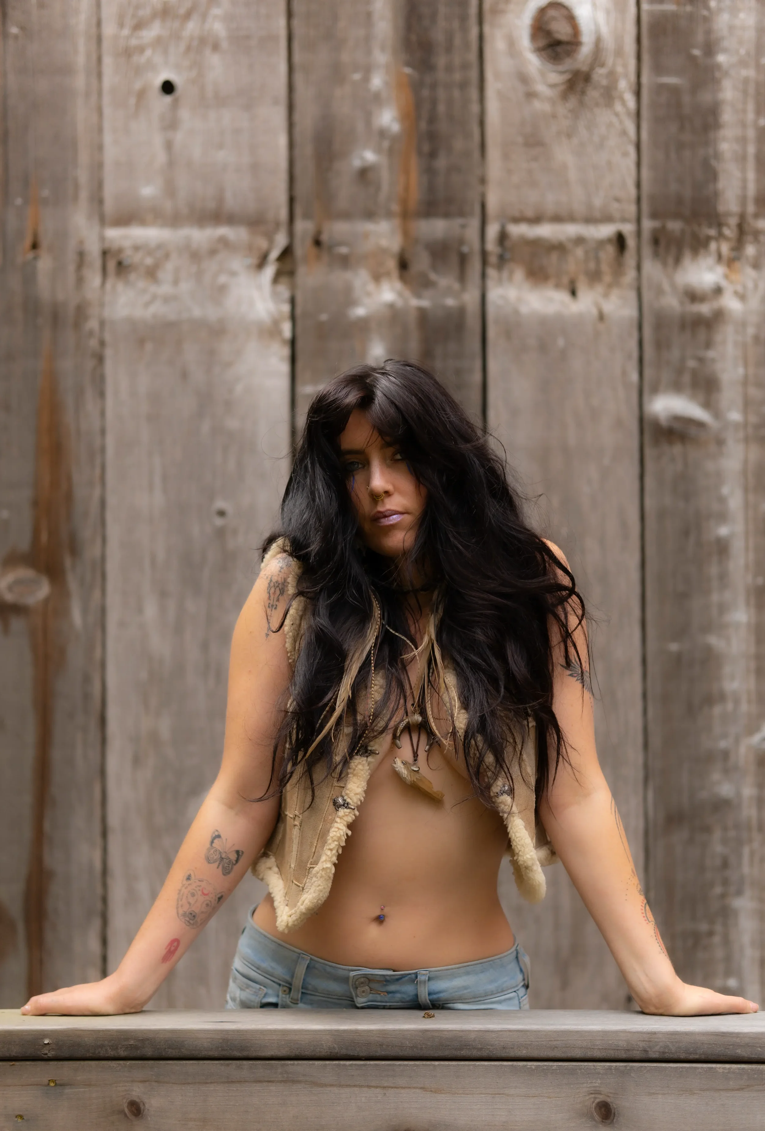 A woman with long dark hair and tattoos on her arms is standing behind a wooden table, leaning forward with her hands resting on the table, against a wooden wall background.