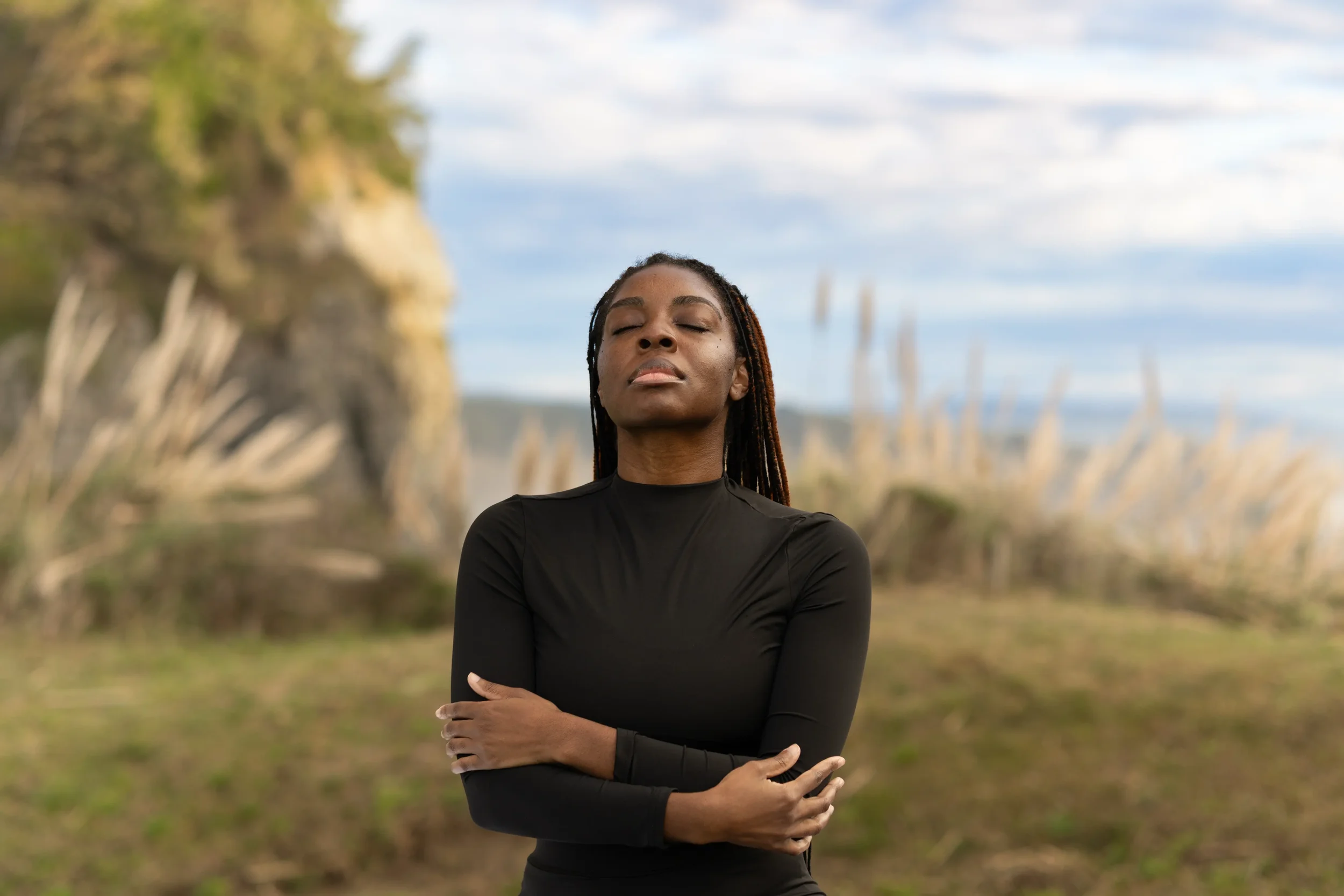 A woman with closed eyes and braided hair poses outdoors with her arms crossed, wearing a black long-sleeved top, and a blurred natural background of trees and sky.