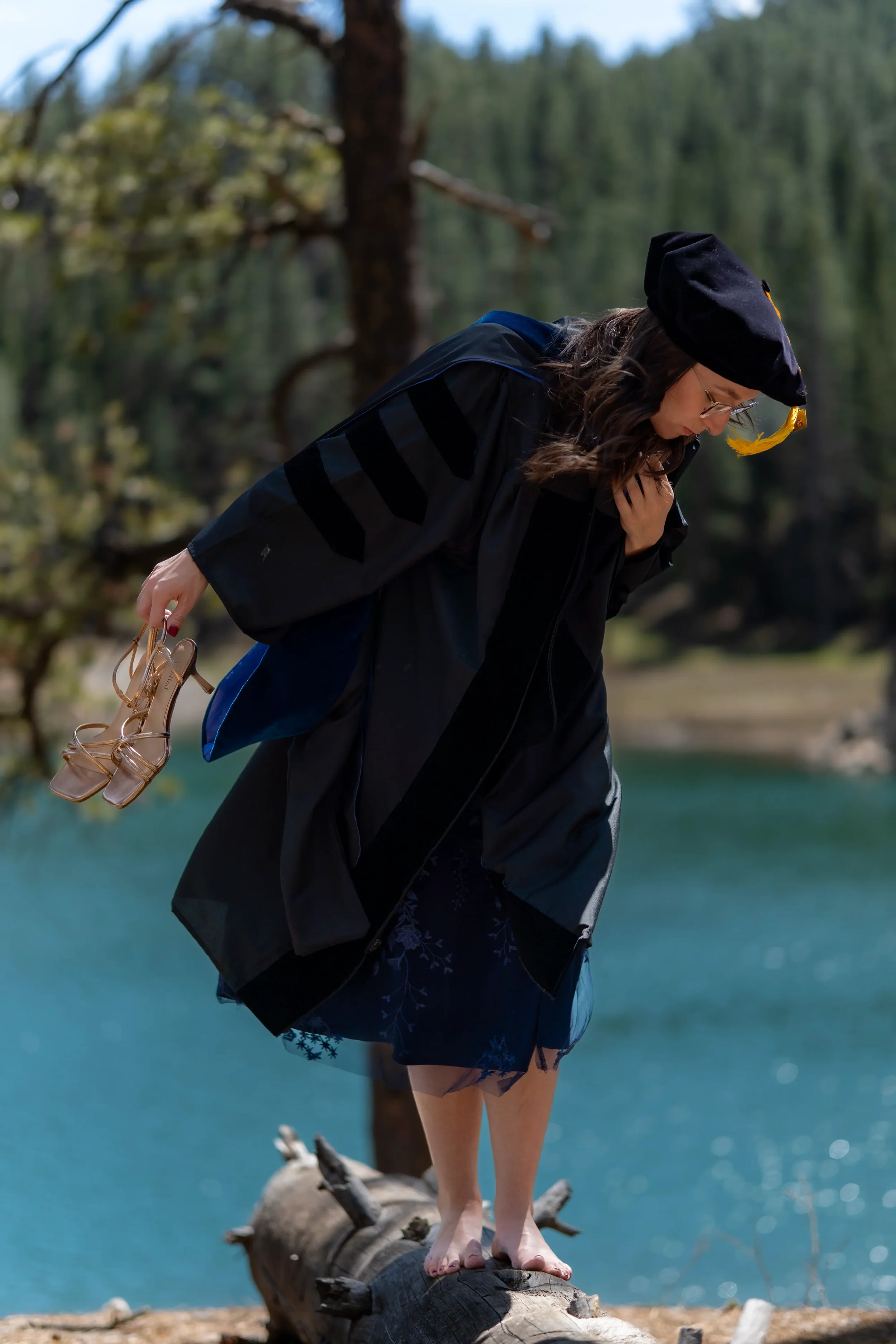 A woman in glasses and a black graduation gown with a cap carrying a pair of beige sandals stands on a fallen tree trunk by a lake, surrounded by trees with a blue sky in the background.