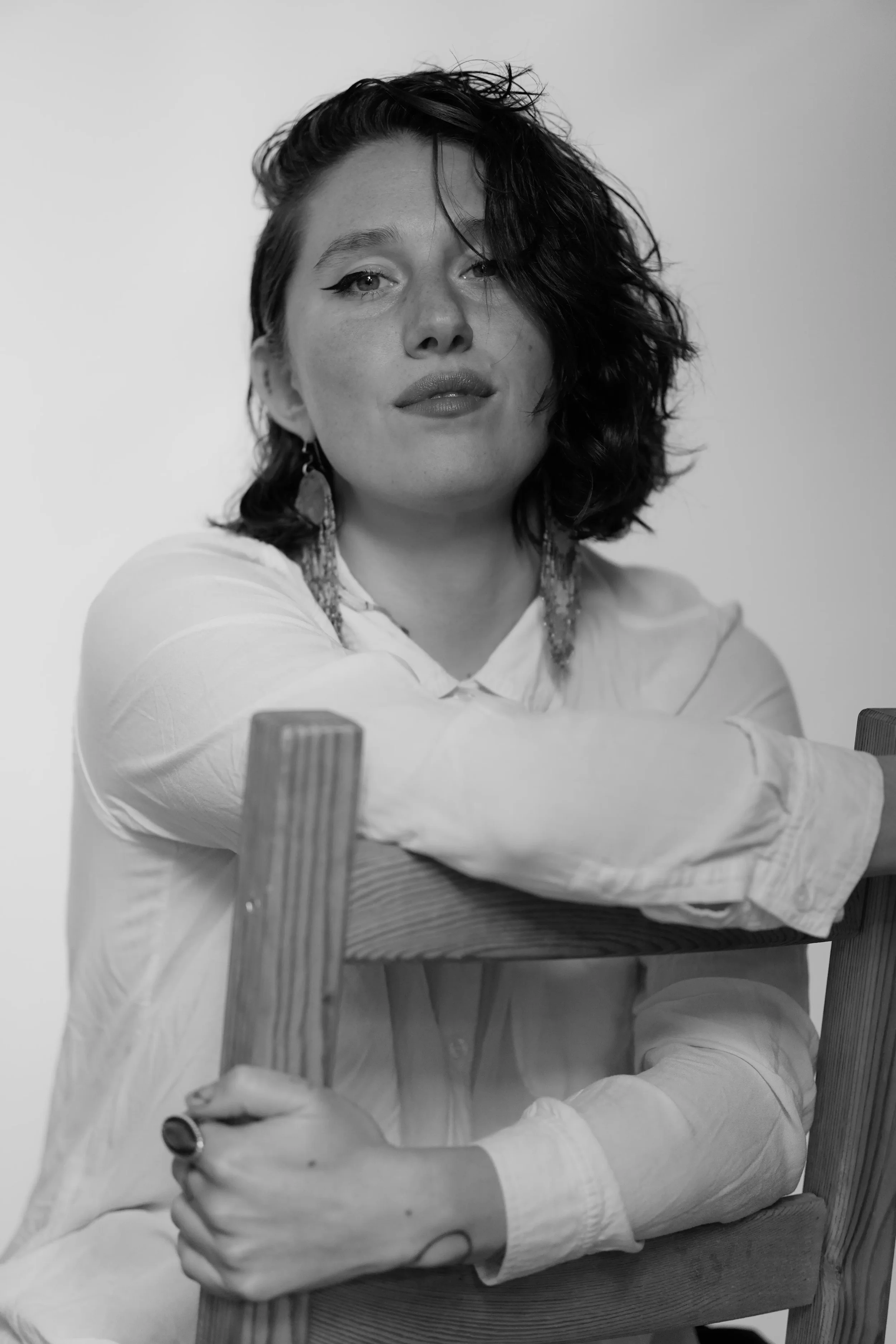 A woman with short, curly hair, wearing a white long-sleeve shirt and earrings, is sitting at a table with her arms resting on the back of a chair.