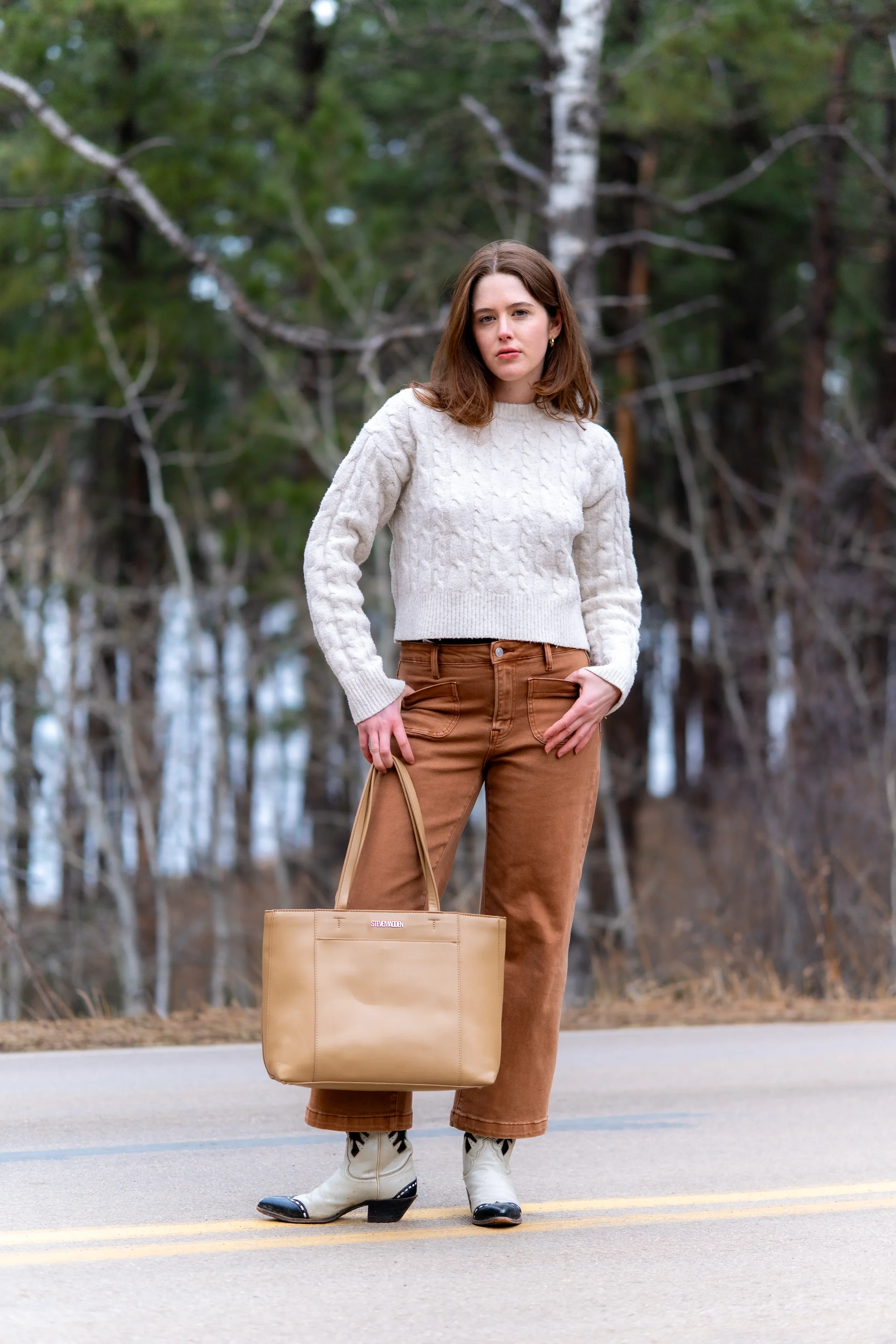A woman with brown hair standing on a road in front of a wooded area, wearing a cream cable-knit sweater, brown pants, and cowboy boots, holding a tan tote bag.
