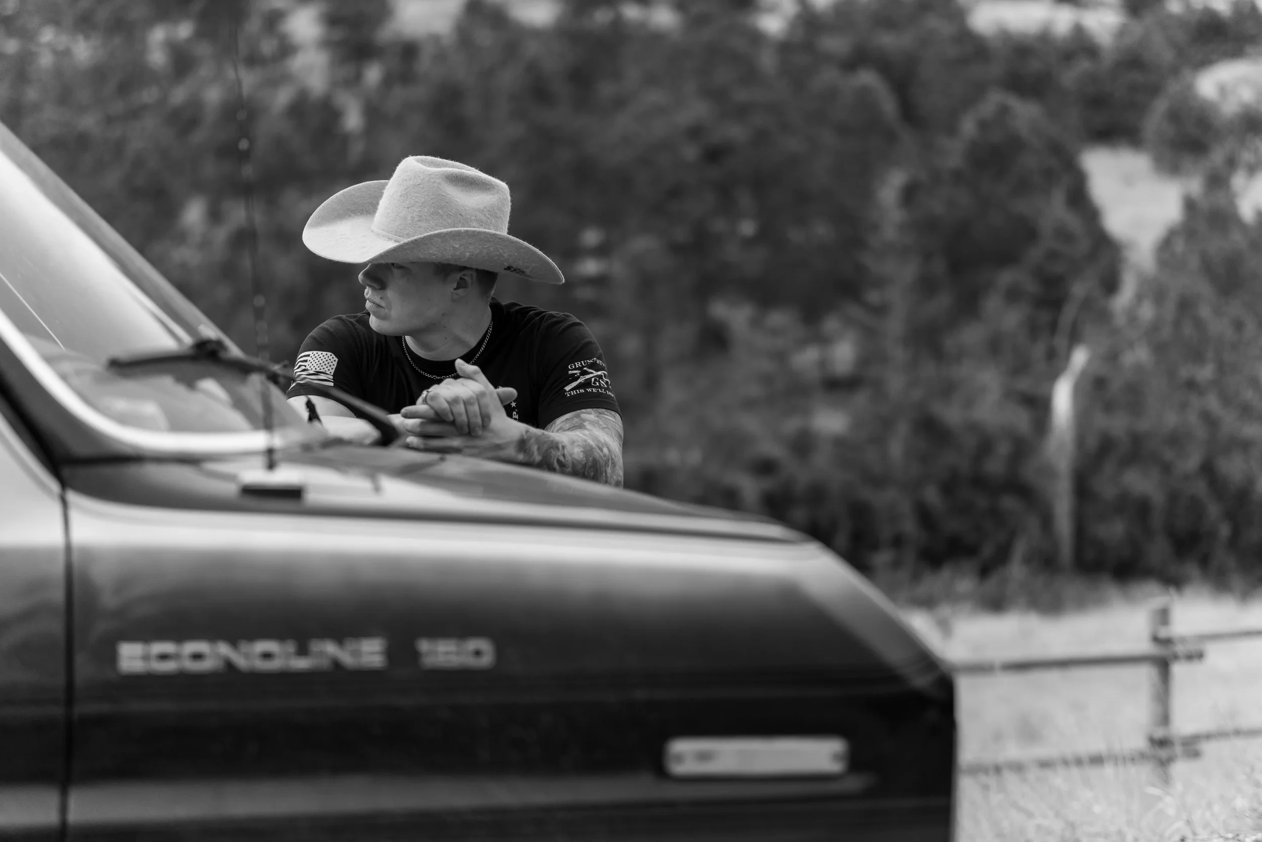 A person wearing a large cowboy hat and black shirt standing beside a police vehicle, with a forested background.