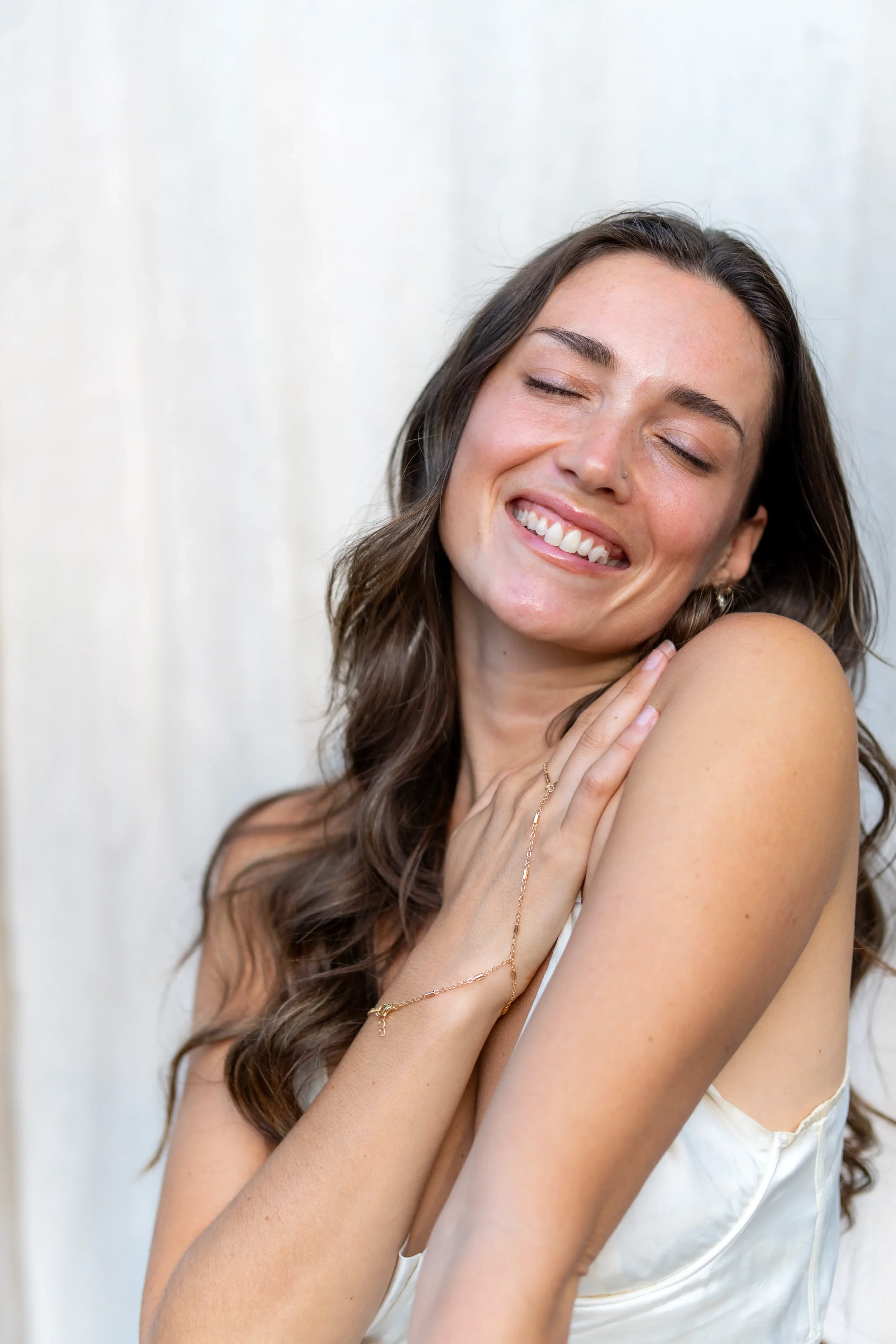 A young woman with long, wavy brown hair smiling with her eyes closed, touching her shoulder, wearing a white slip dress and a delicate gold bracelet, against a plain white background.