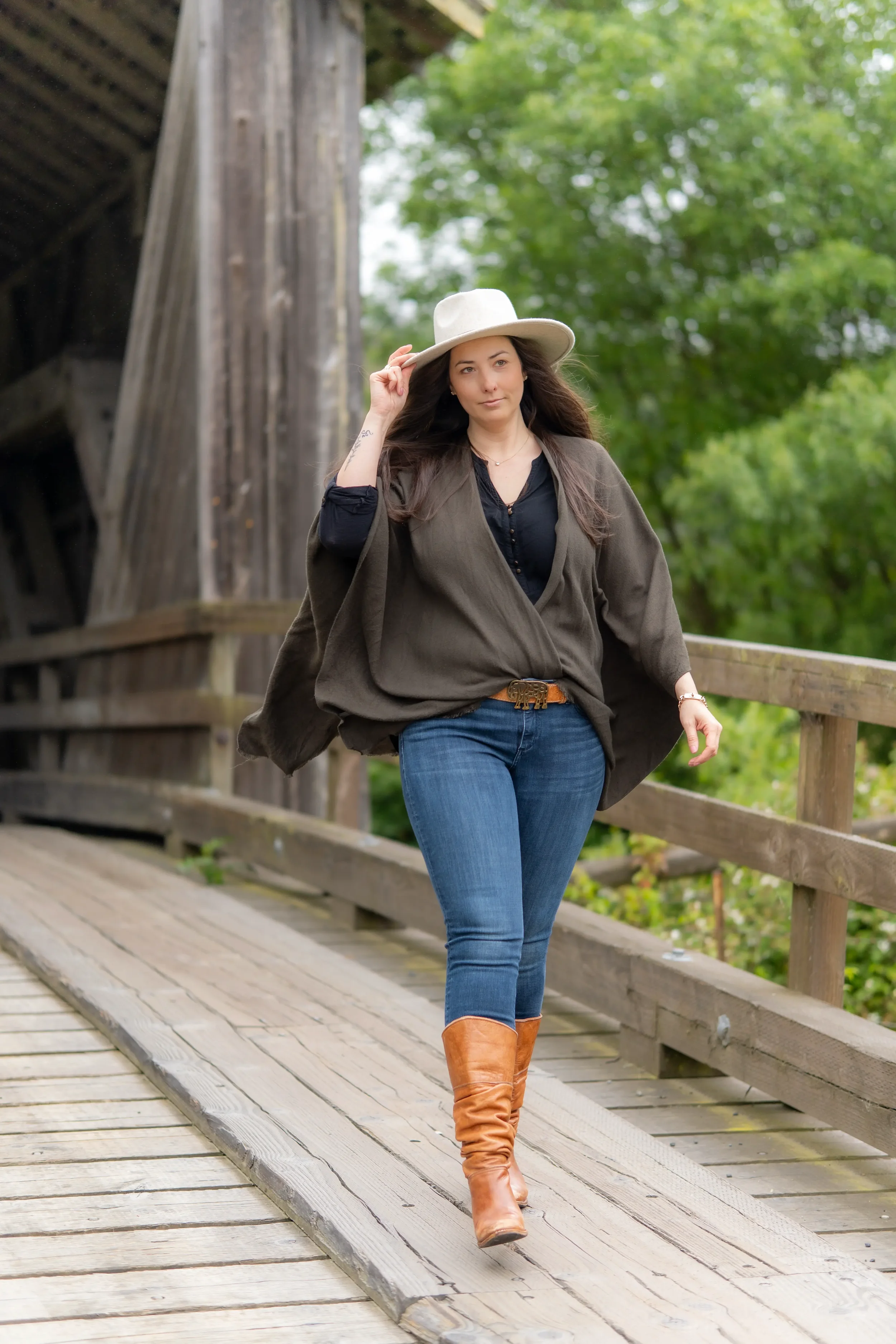 A woman walking on a wooden bridge, wearing a wide-brimmed hat, dark blouse, brown cape, blue jeans, and knee-high tan boots, with a green park background.