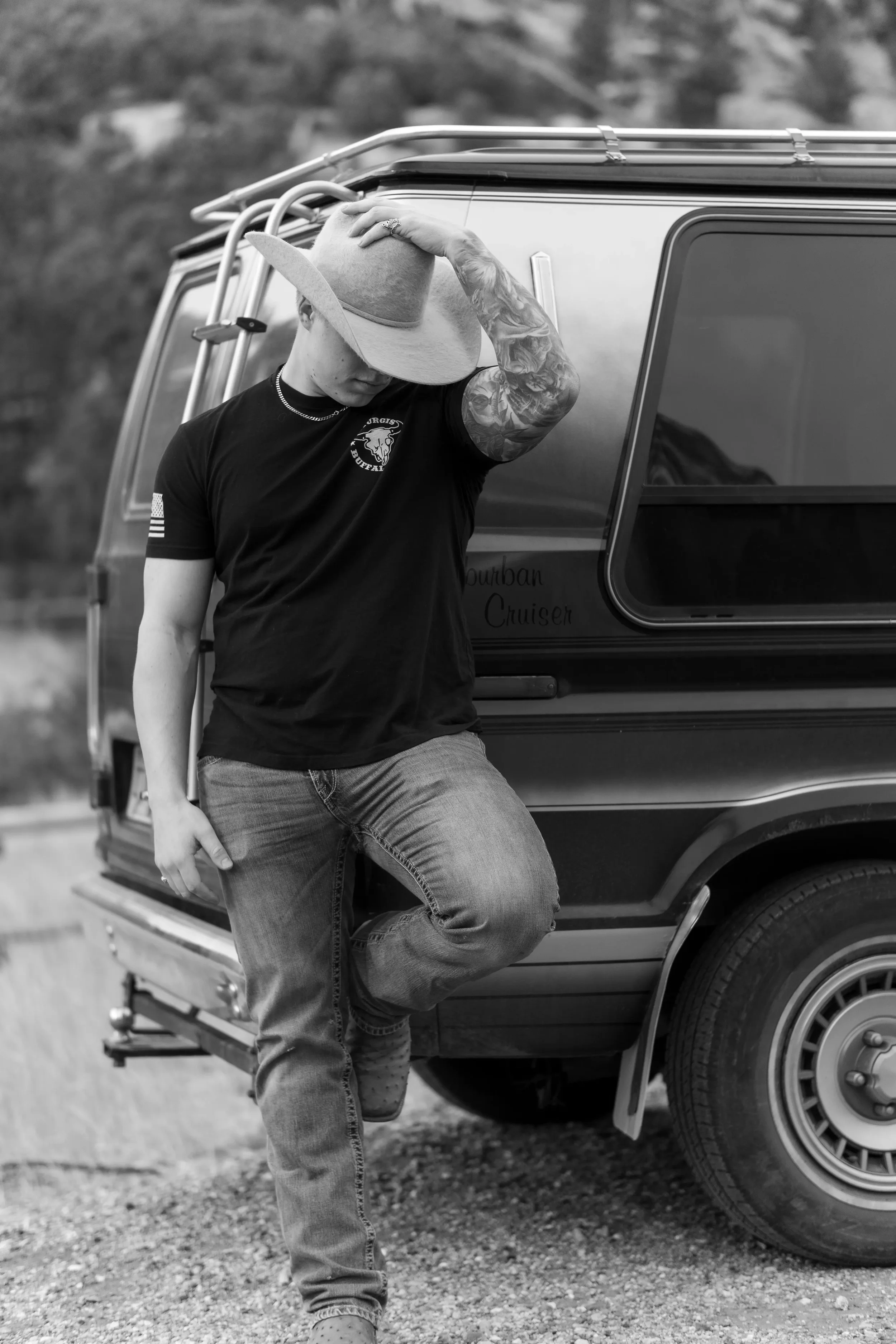 A person with tattoos and a side-shaved hairstyle leaning against a vintage camper van with a logo on the door that reads 'Durban Cruiser' while holding a wide-brimmed hat and looking down, in black and white. The person is wearing a dark t-shirt, je
