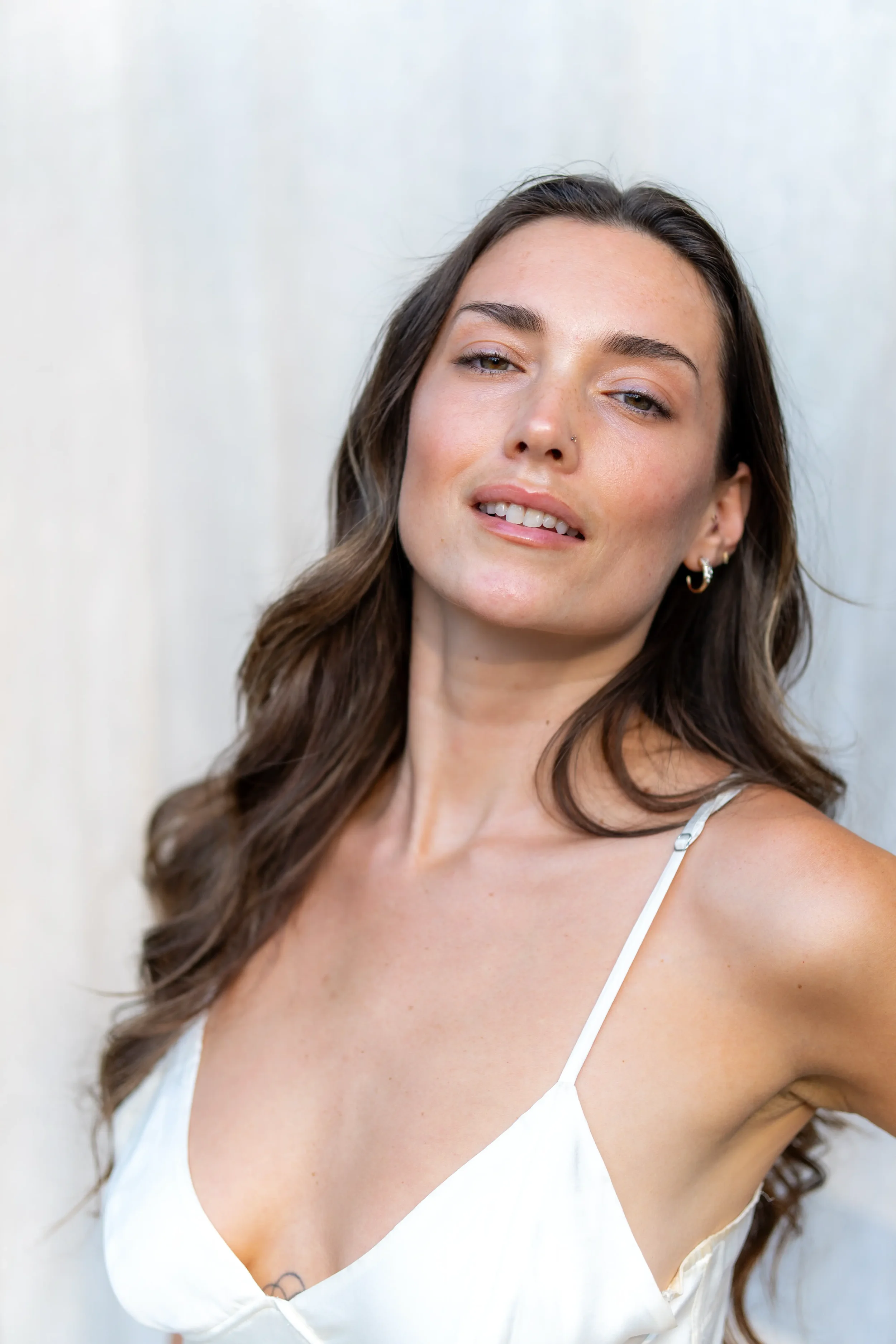 Close-up of a woman with long brown hair and light lip color, wearing a white spaghetti-strap top with a tattoo visible on her chest, against a plain light background.