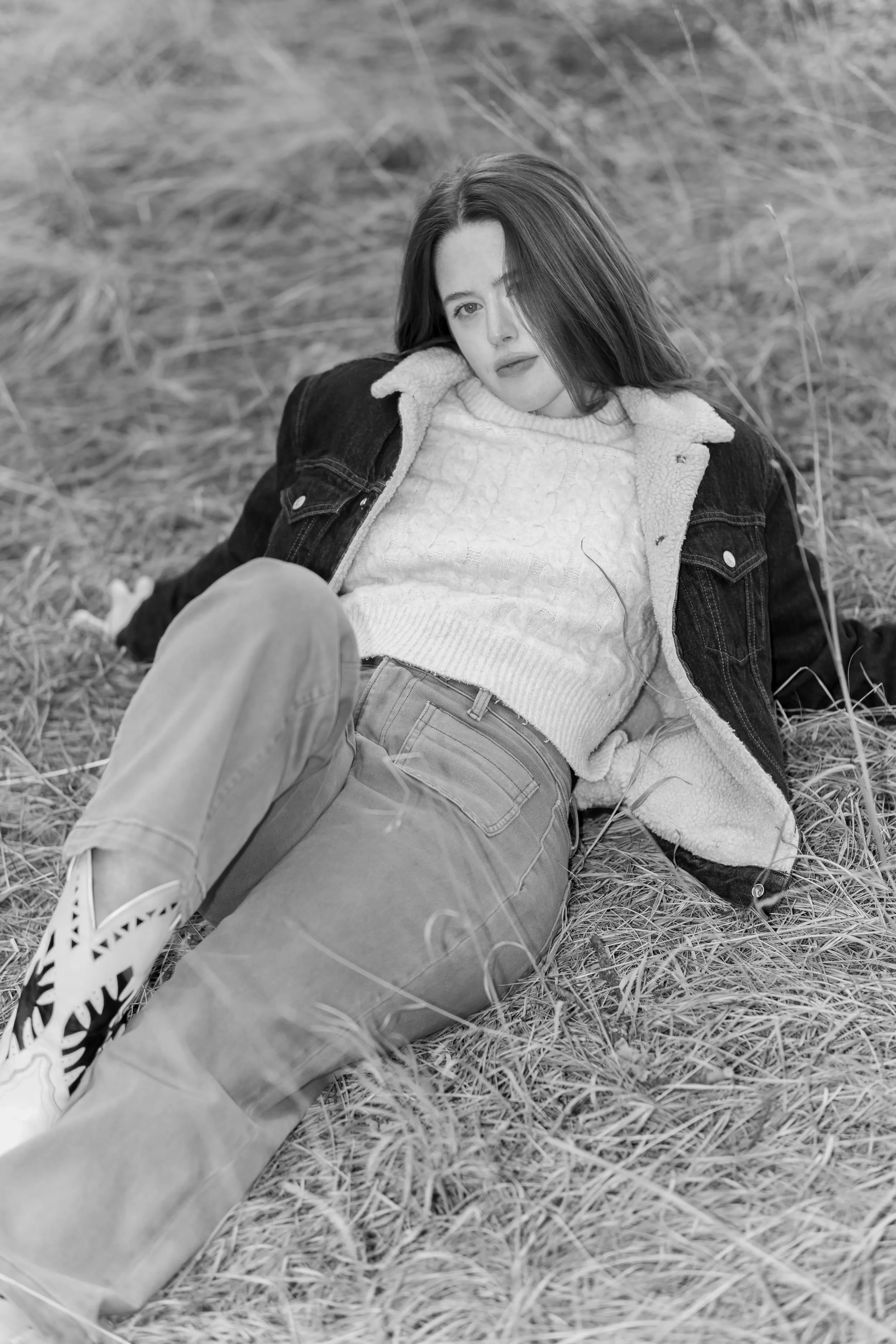 A woman reclining on grass, wearing a white sweater and a dark jacket, with her head resting on her hand, looking at the camera.
