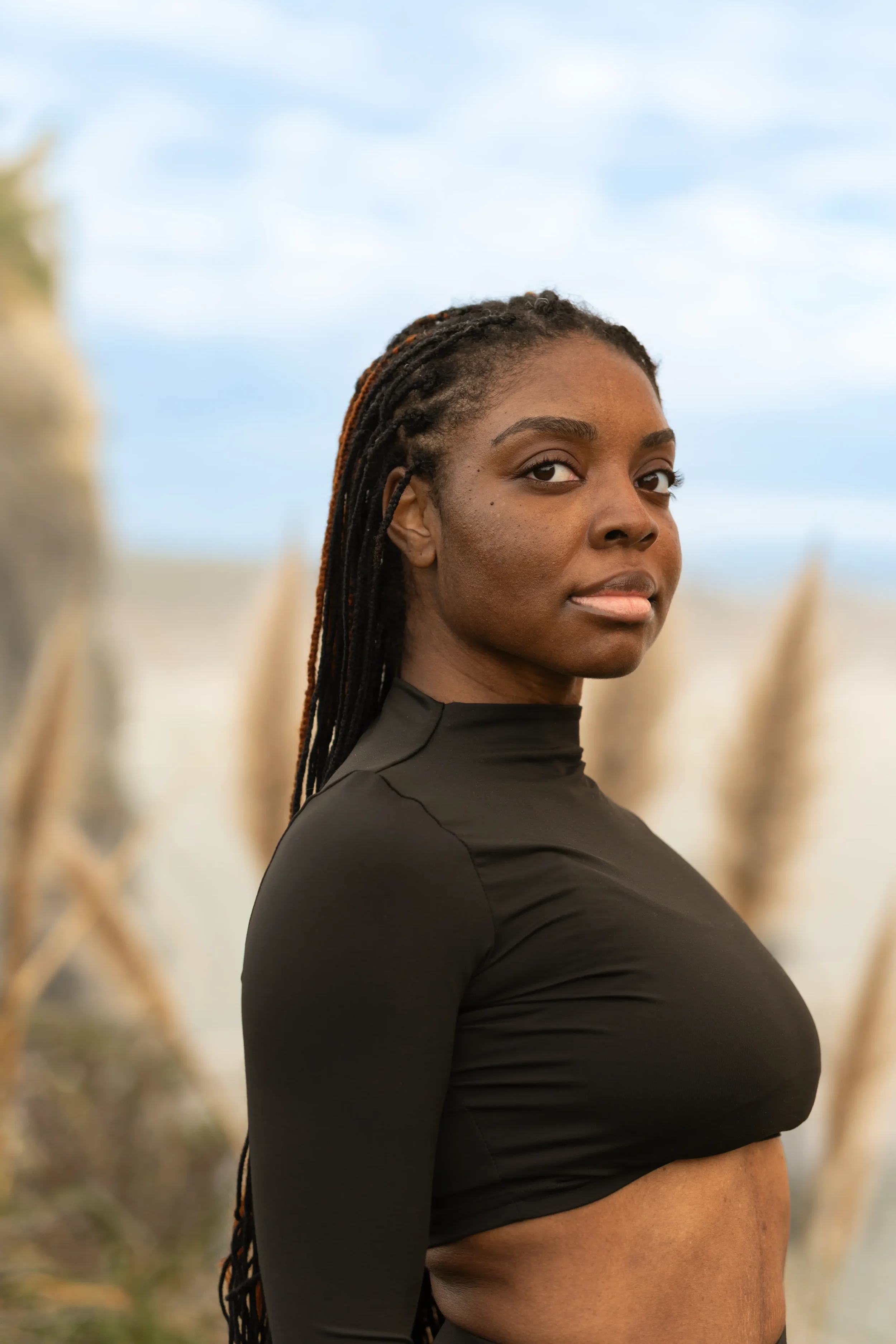 A woman with dark skin and dreadlocks stands outdoors among blurred tall yellow grass under a cloudy sky, wearing a black long-sleeved top and looking at the camera.