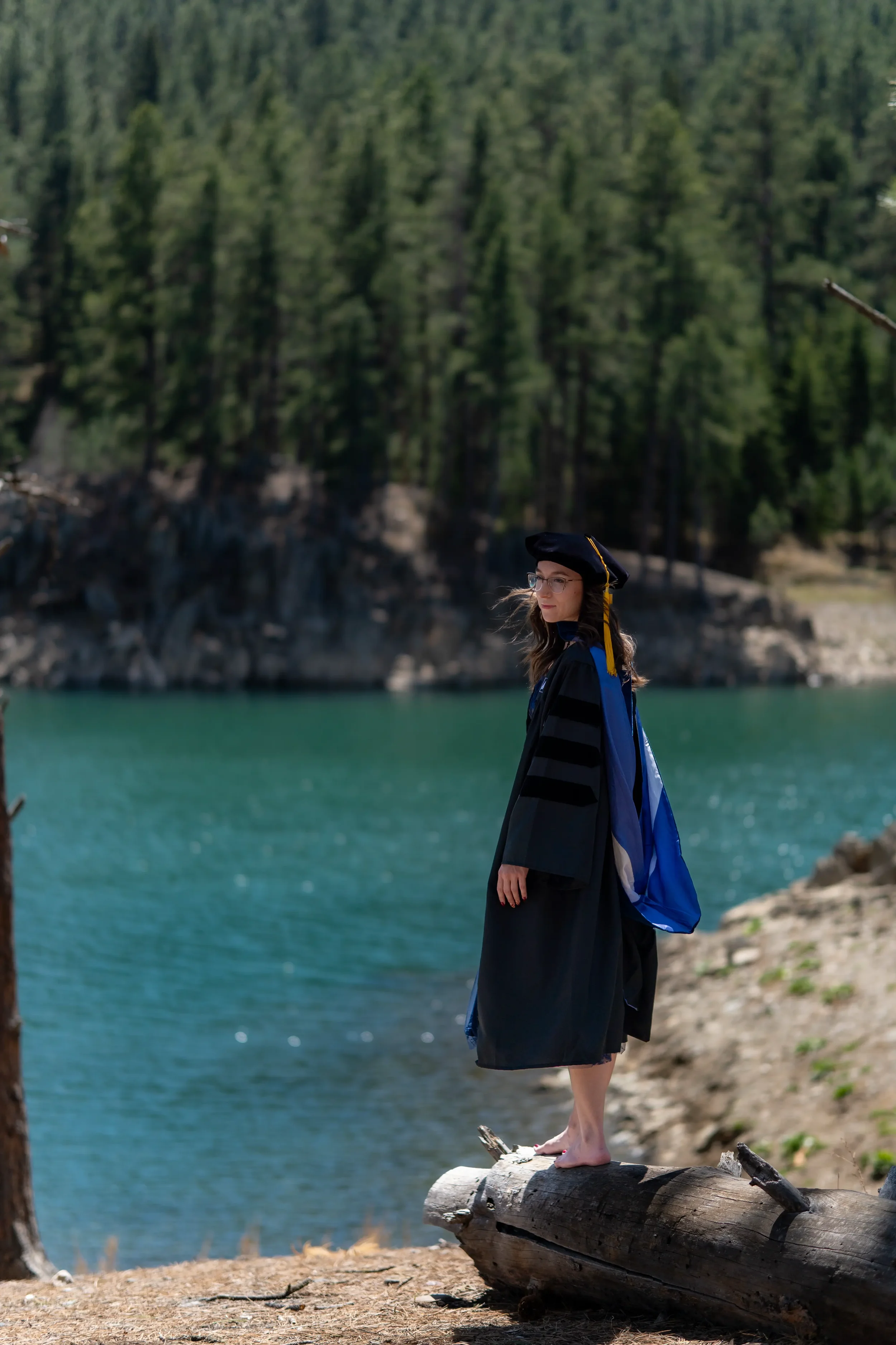 A woman in a graduation gown and cap standing on a log by a lake, with a forested hillside in the background.