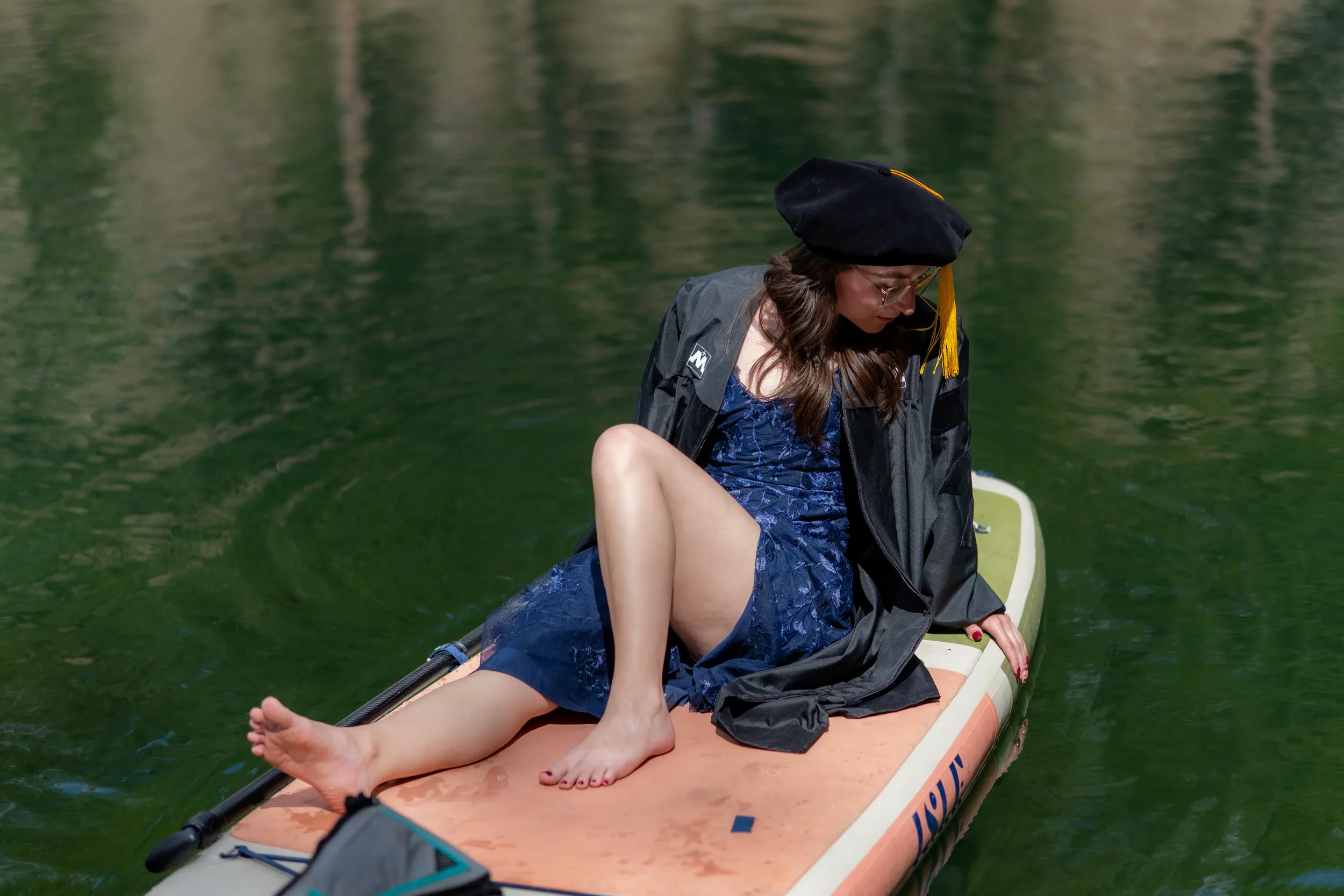 A woman in a blue dress sits on a paddleboard on a green body of water, with her legs extended and a black jacket draped over her shoulders. She wears glasses and a black graduation cap with a yellow tassel.