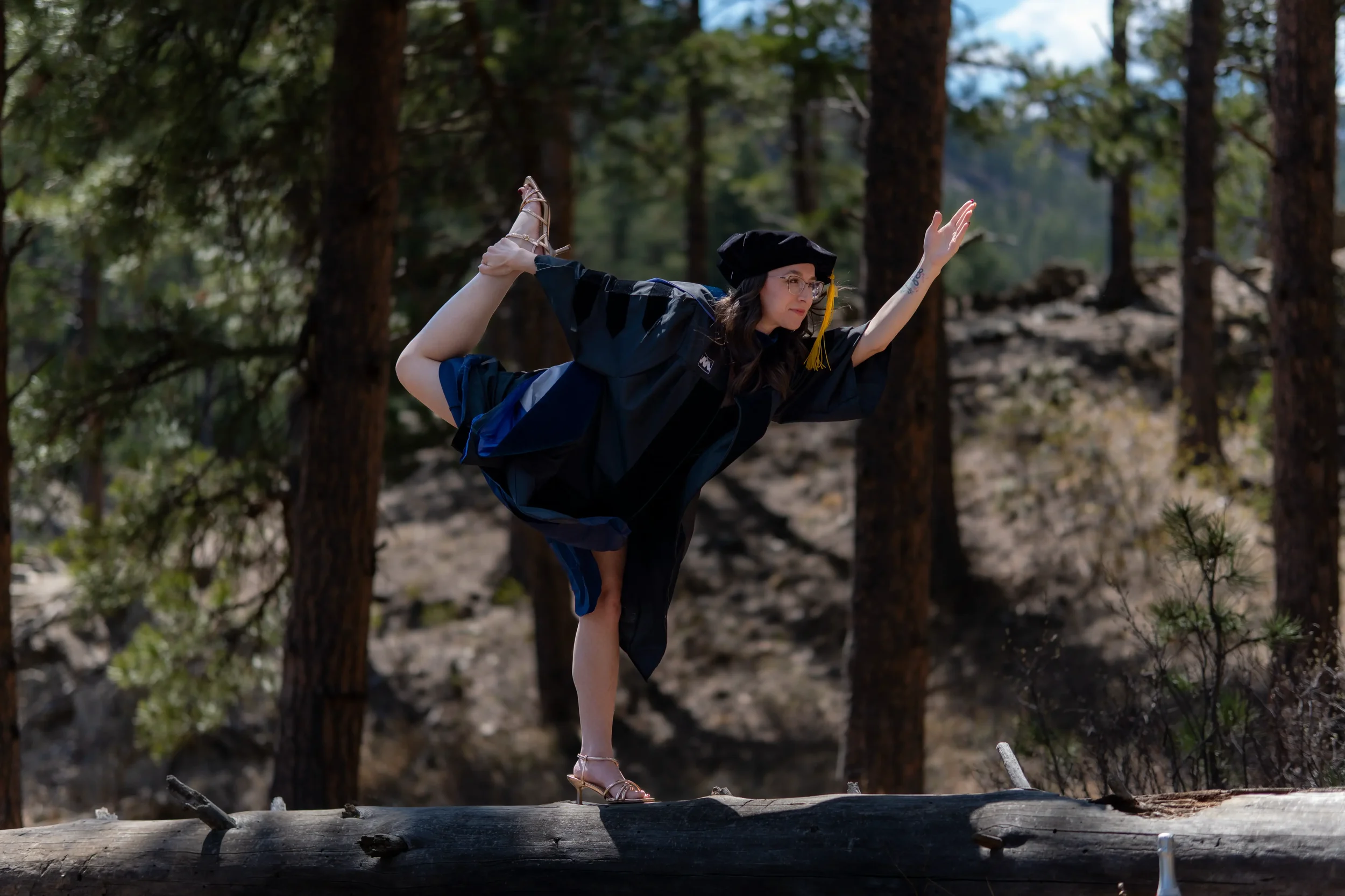 Woman in a graduation cap and gown performing a yoga pose on a fallen tree in a forest.