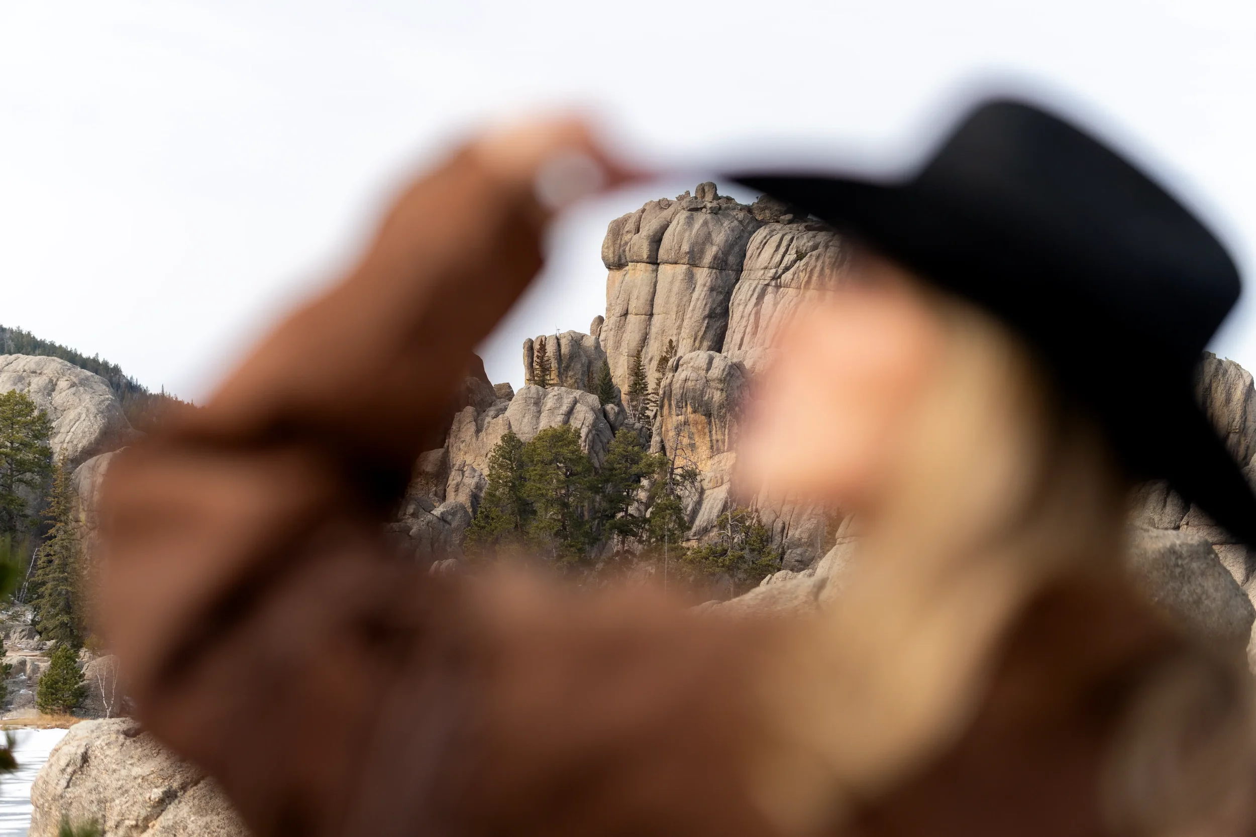 A hand holding a black camera capturing a photo of granite rock formations and pine trees in a mountainous landscape.