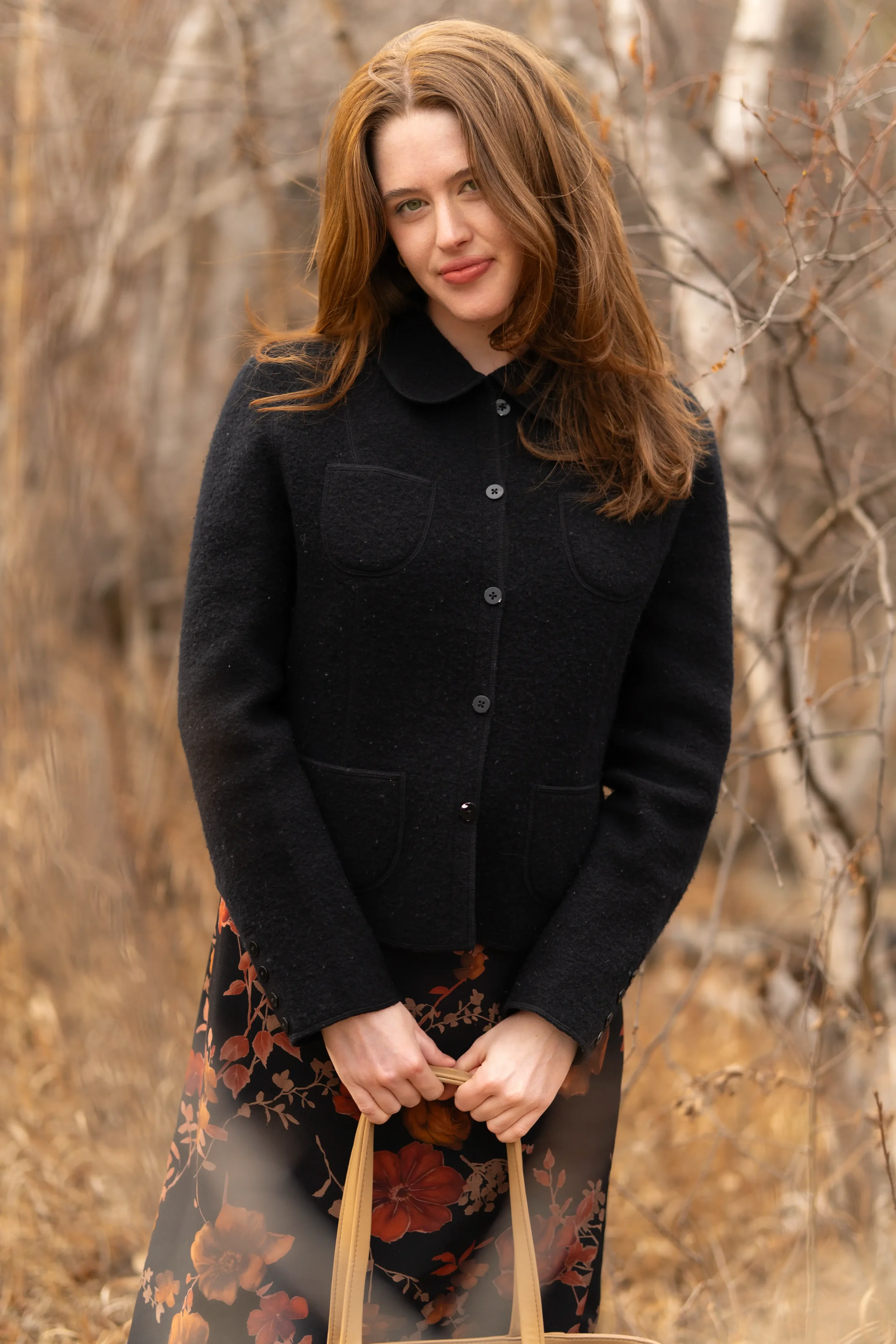 A woman with long red hair wearing a black coat stands outdoors in a wooded area with leafless trees. She is holding a bag with a floral design featuring large red, orange, and brown flowers.