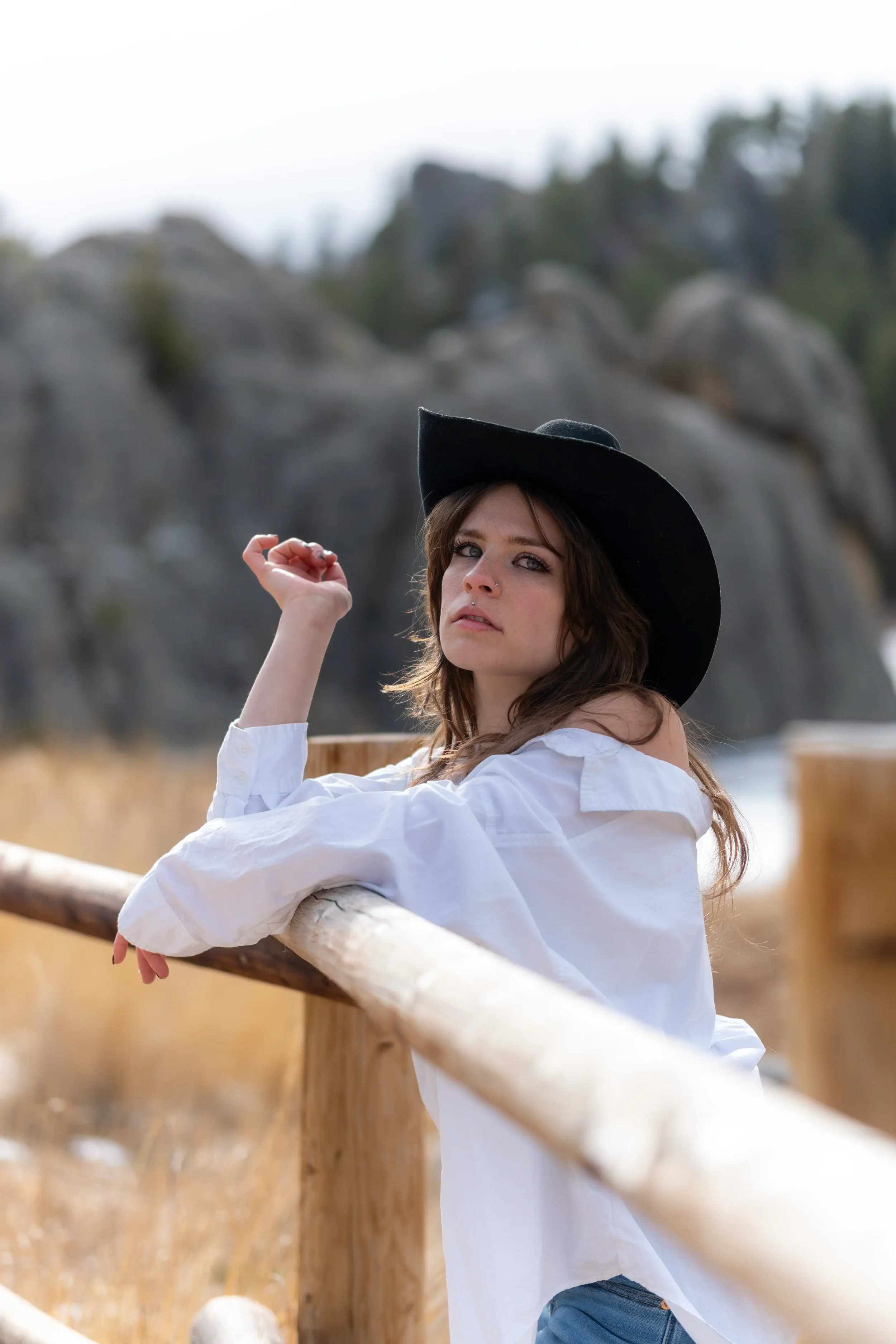 A young woman with wavy brown hair, wearing a black wide-brimmed hat and a white off-the-shoulder blouse, is sitting or leaning on a wooden railing outdoors. She has a relaxed expression and is looking at the camera with a slight expression of intrig