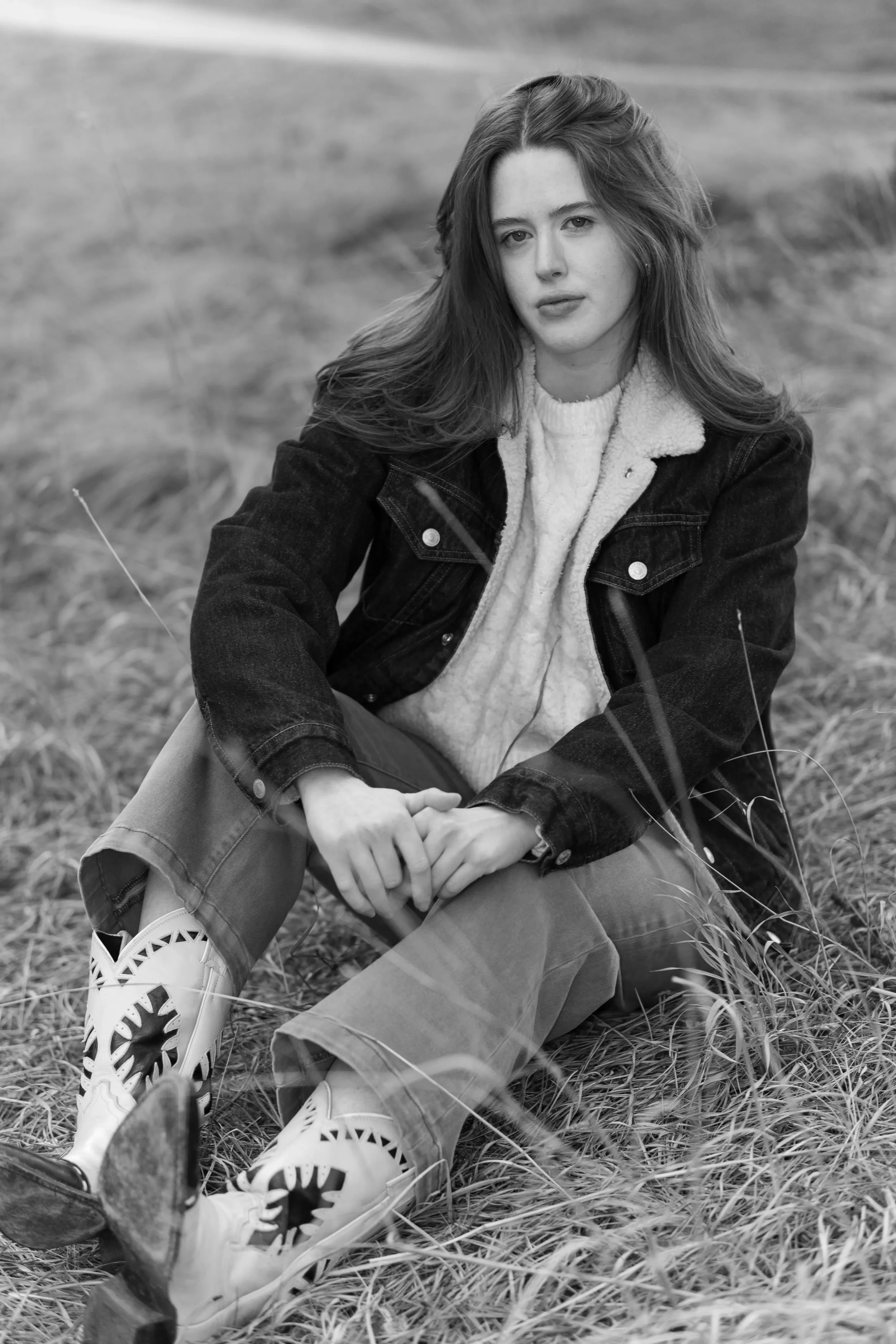 A young woman with long hair sitting outdoors on grass, wearing a jacket, sweater, and patterned shoes, looking at the camera.