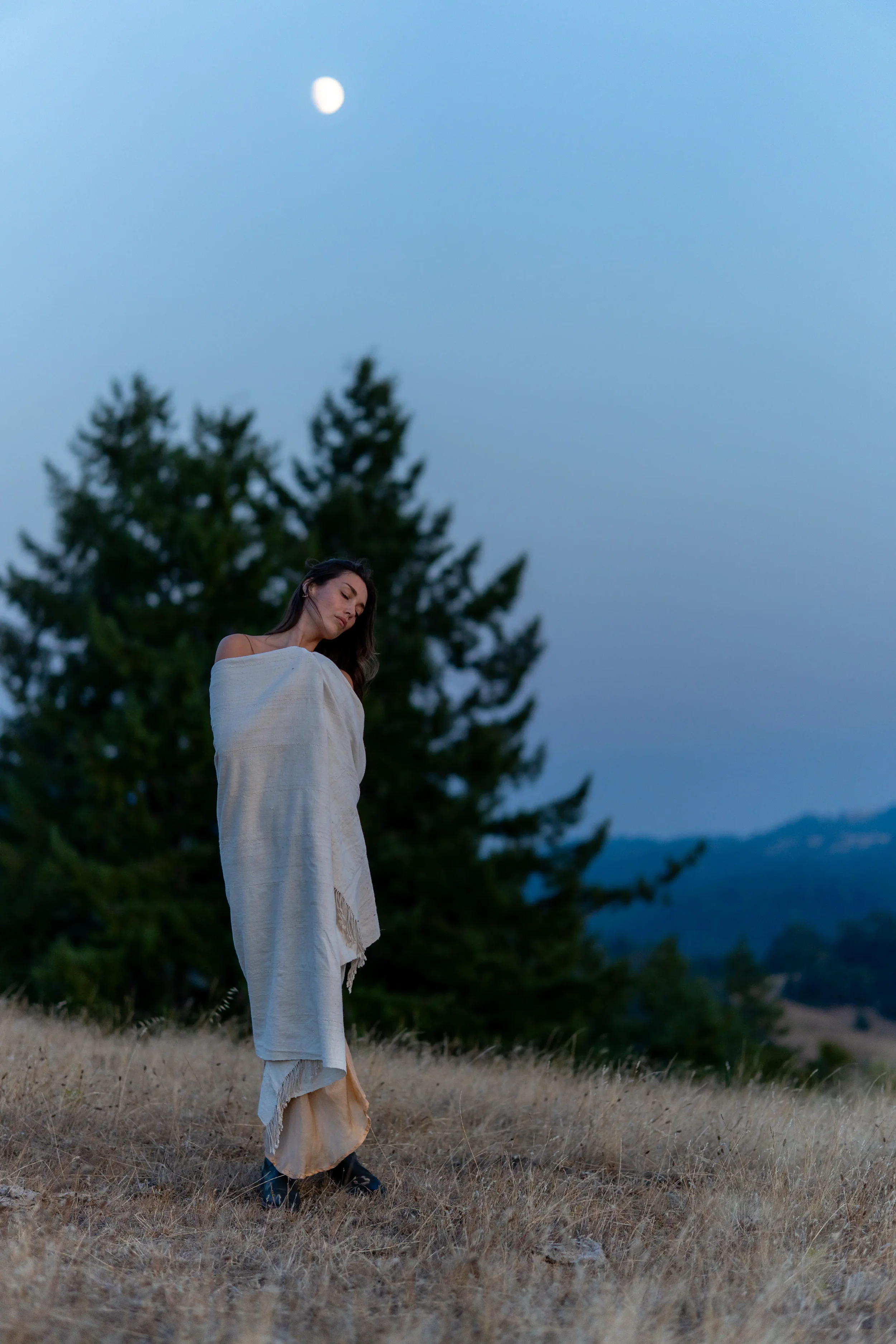 A woman standing outdoors in a field at dusk, wrapped in a white blanket, with a large evergreen tree in the background and the moon visible in the sky.