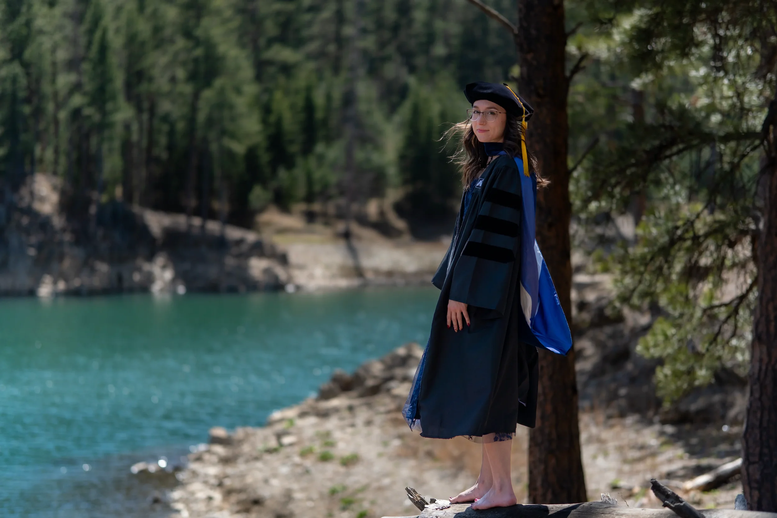A woman in a graduation cap and gown standing on a fallen log by a lake with trees in the background.