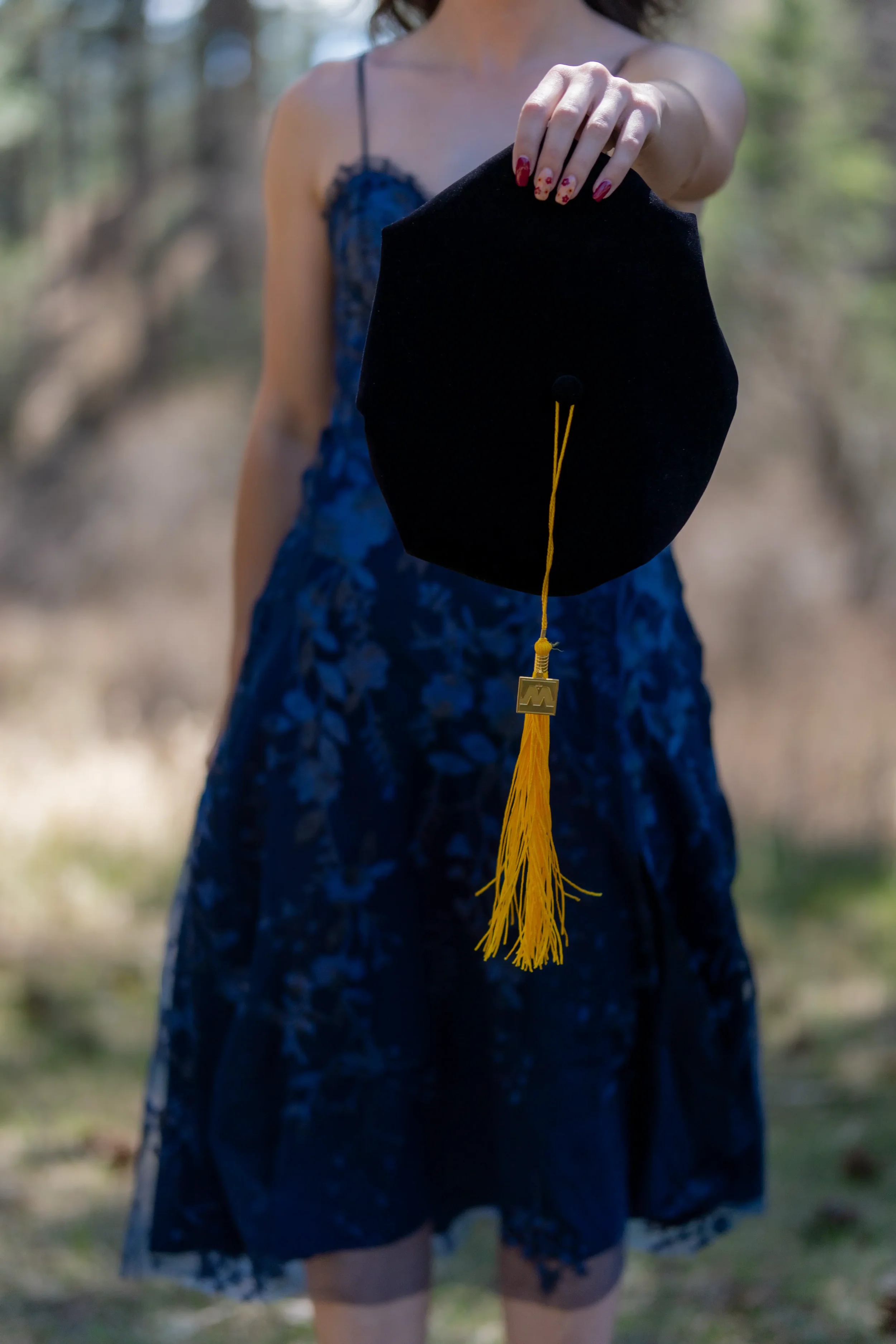 A woman in a blue floral dress holding a black graduation cap with a yellow tassel outdoors.