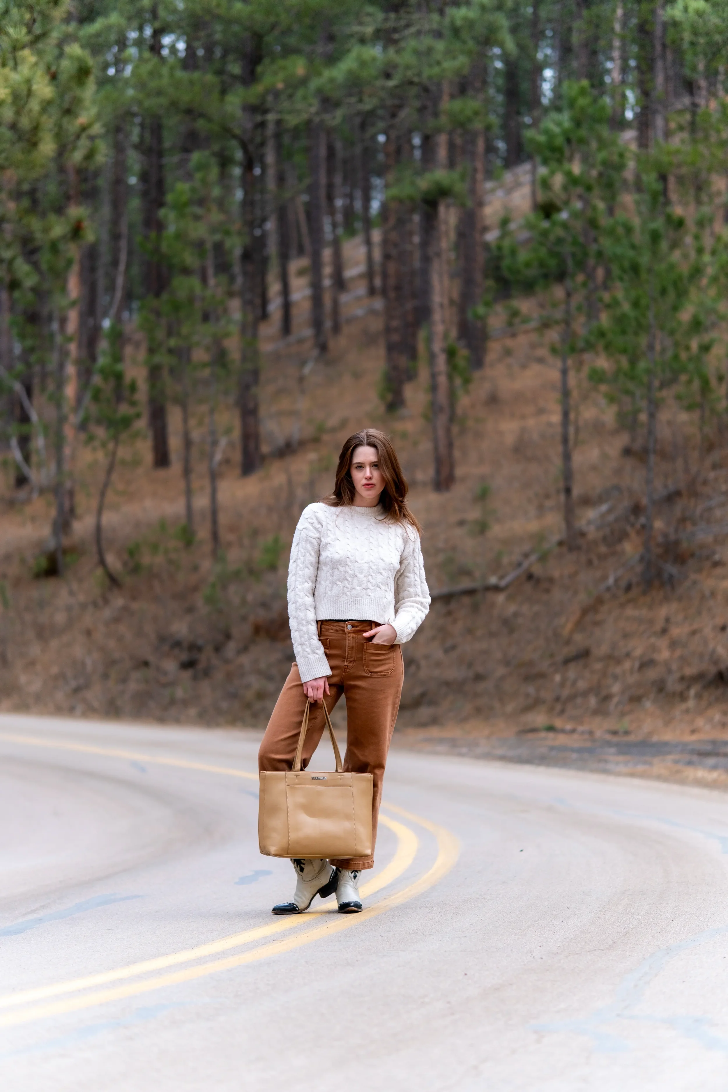 A woman with long brown hair dressed in a white sweater and brown pants standing on a rural road with yellow lines, holding a beige tote bag, with pine trees and a hill in the background.