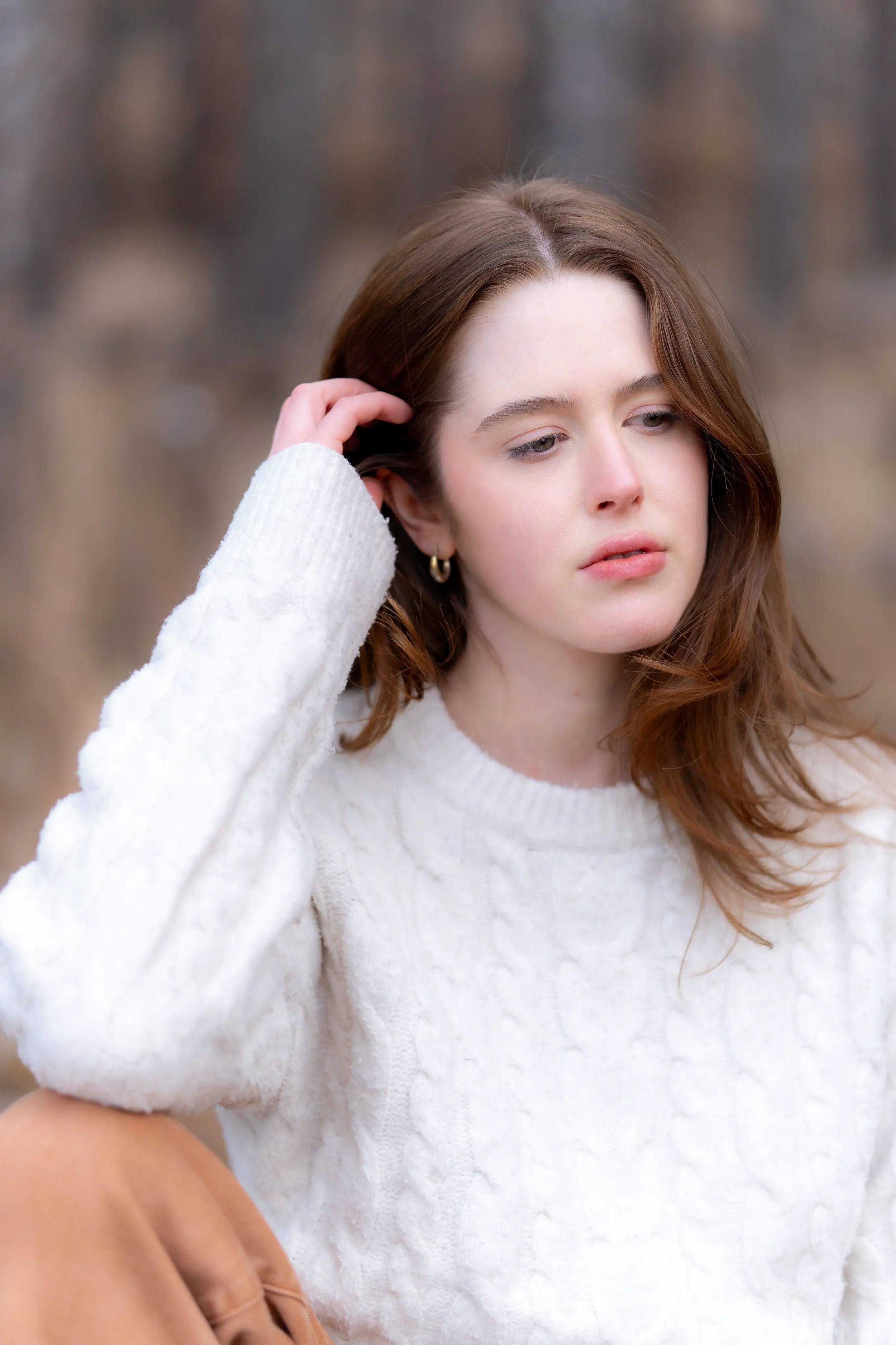 A young woman with light skin and long, wavy, reddish-brown hair is wearing a white knit sweater and small gold hoop earrings. She is outdoors, touching her hair with her right hand, with a neutral facial expression and a blurred natural background.