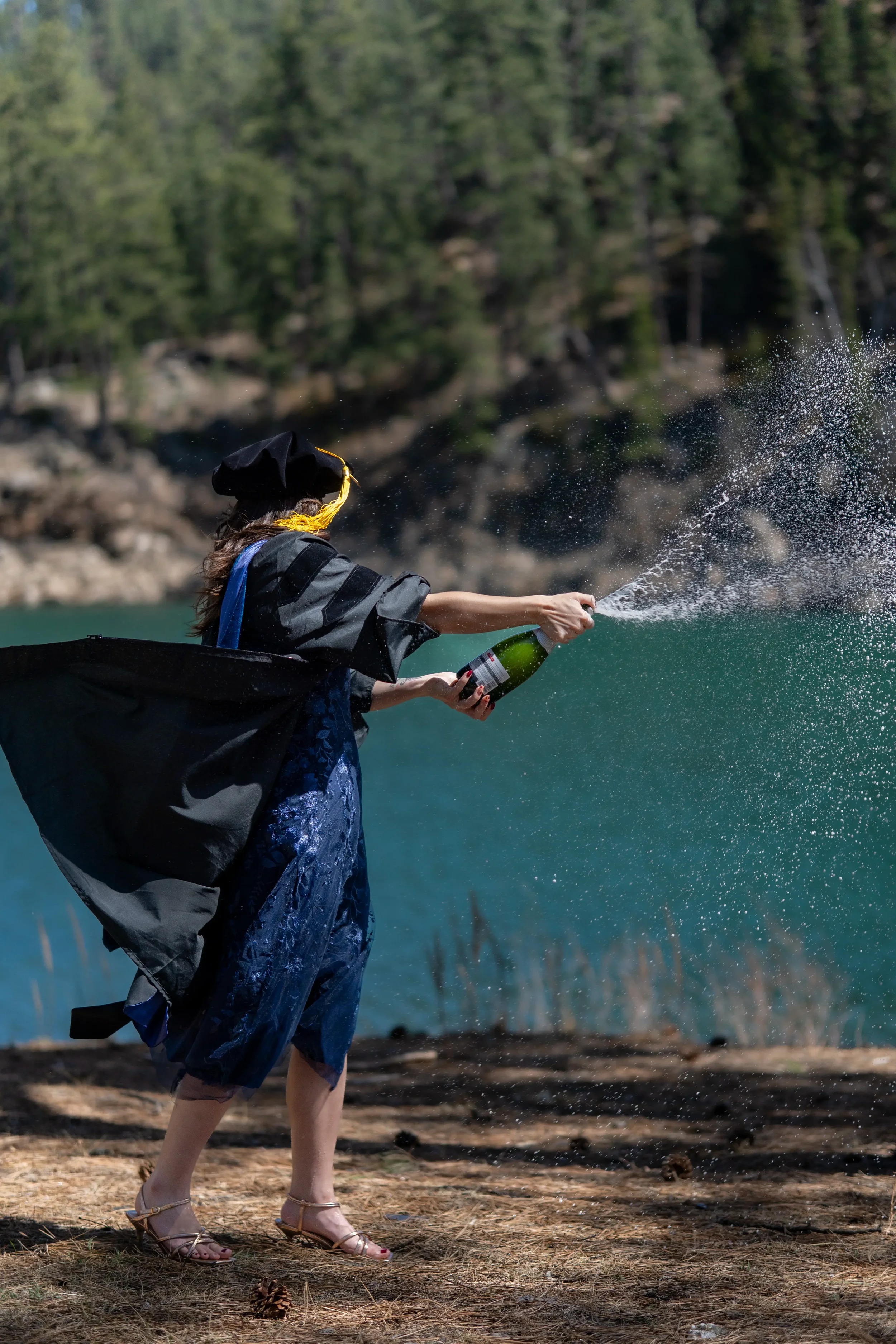 A woman in a graduation cap and gown opening a bottle of champagne outdoors near a body of water, with trees in the background.