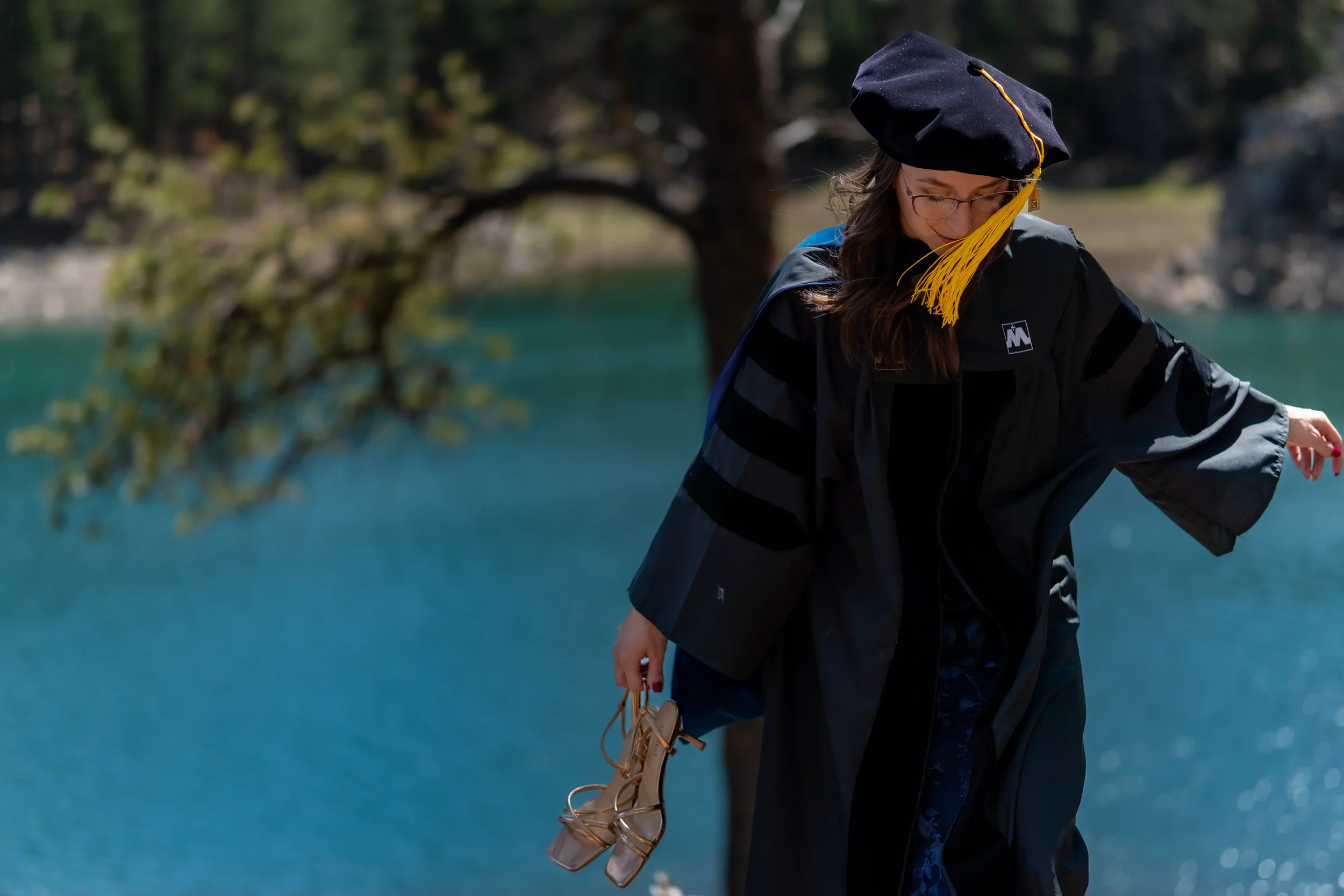 A person wearing a graduation gown and cap, holding a pair of tan high-heeled shoes, standing outdoors near a body of water with trees in the background.