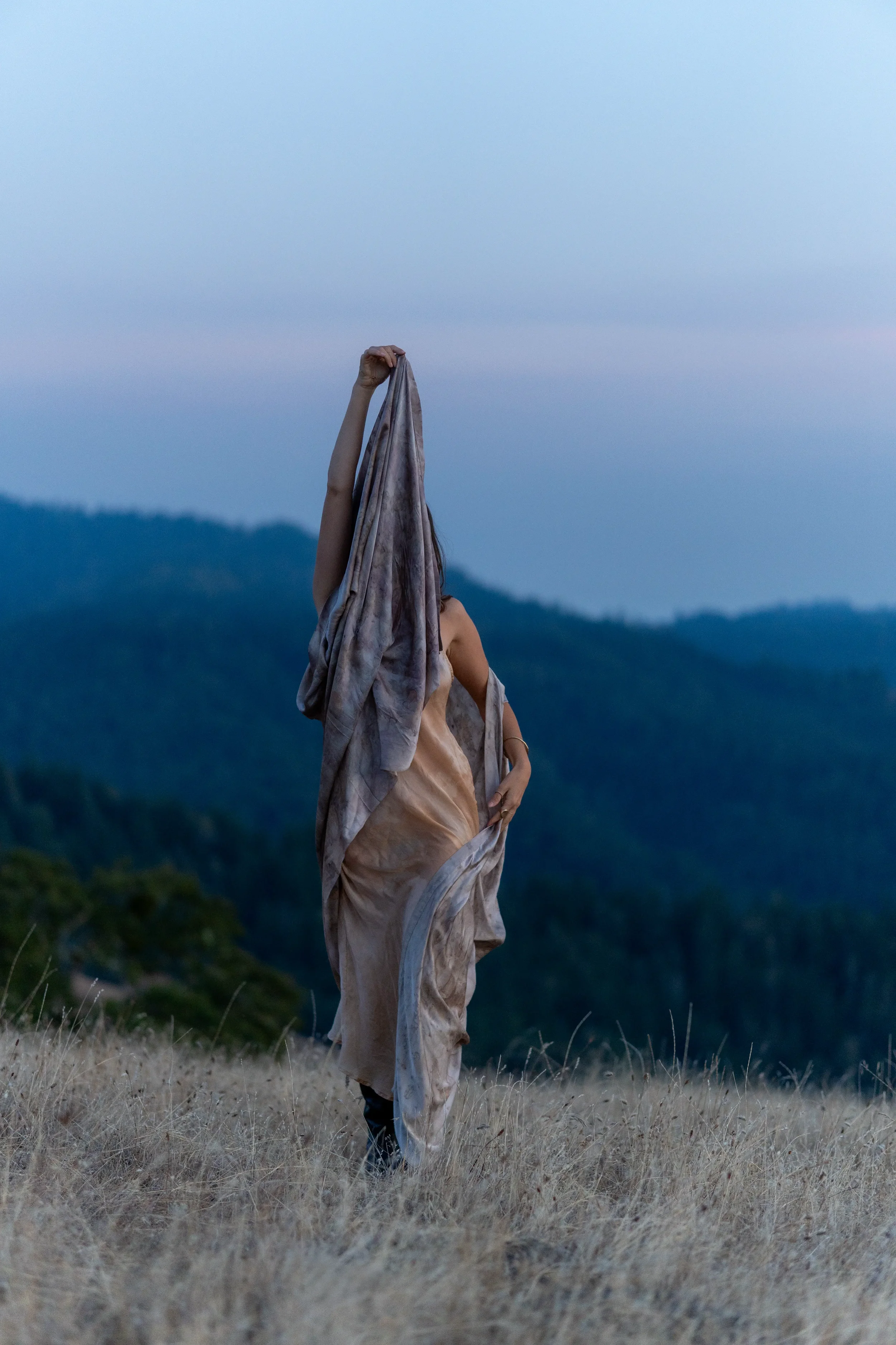 Person holding up a piece of cloth or fabric outdoors in a grassy, hilly landscape during dusk or dawn.