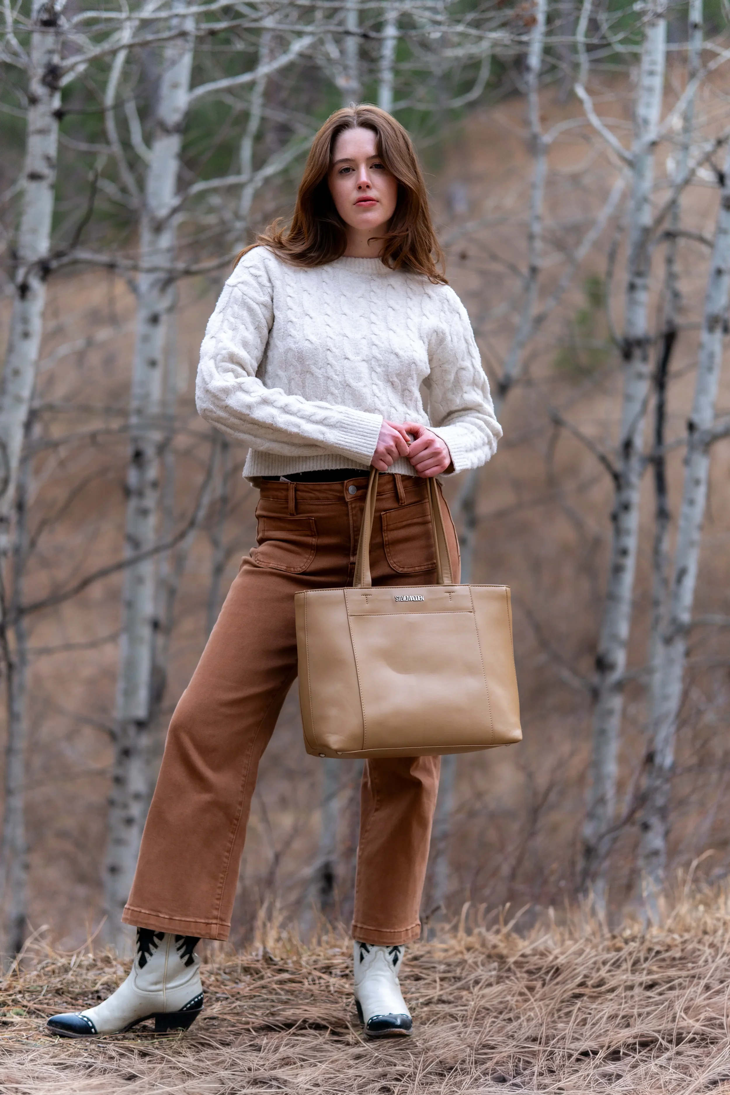 A woman stands outdoors in a forested area with leafless trees, wearing a white sweater, brown pants, and white cowboy boots, holding a beige tote bag.