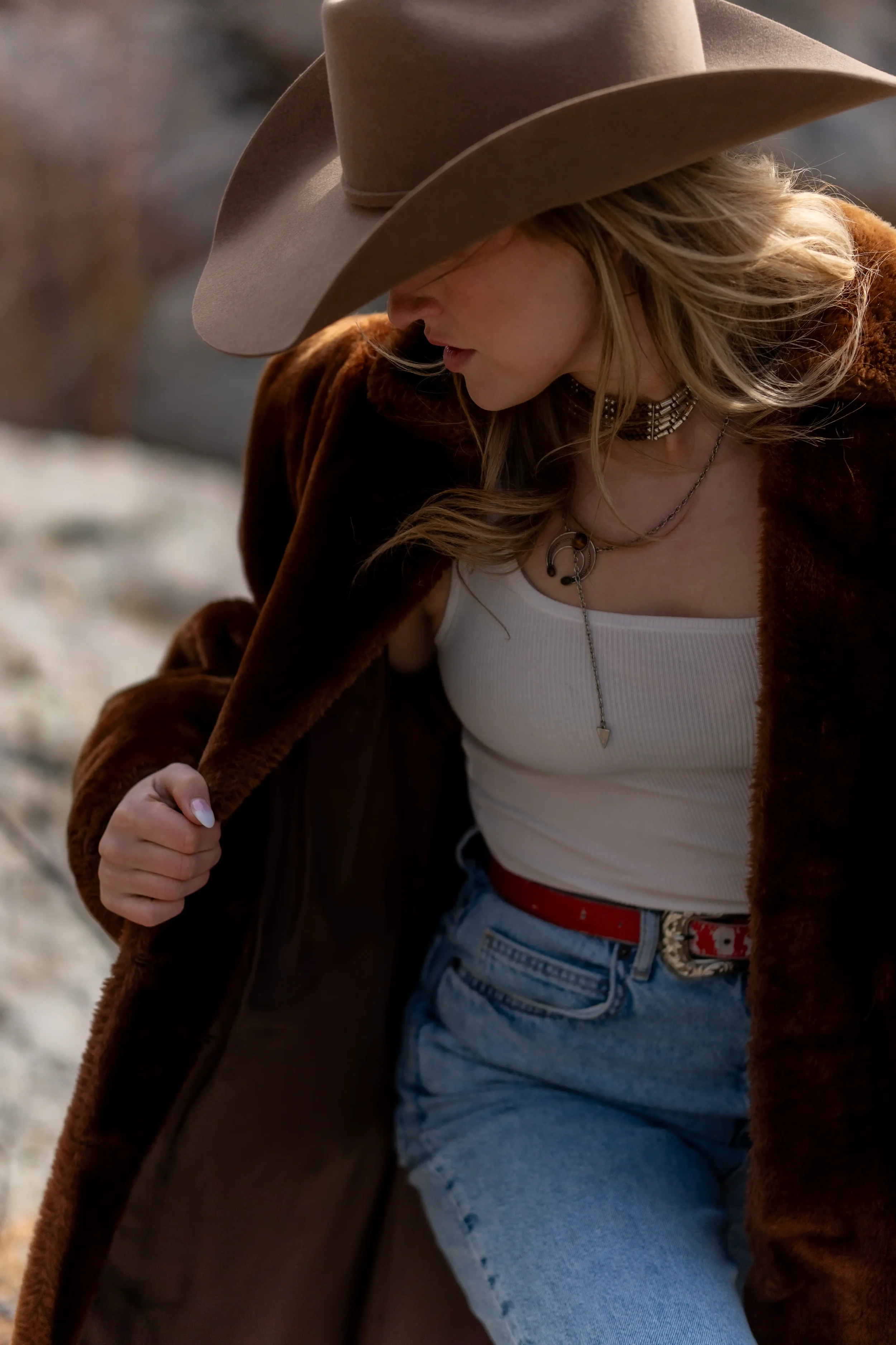 A young woman wearing a beige cowboy hat, a dark brown jacket, a white tank top, and jeans, with layered necklaces, outdoors during daytime.