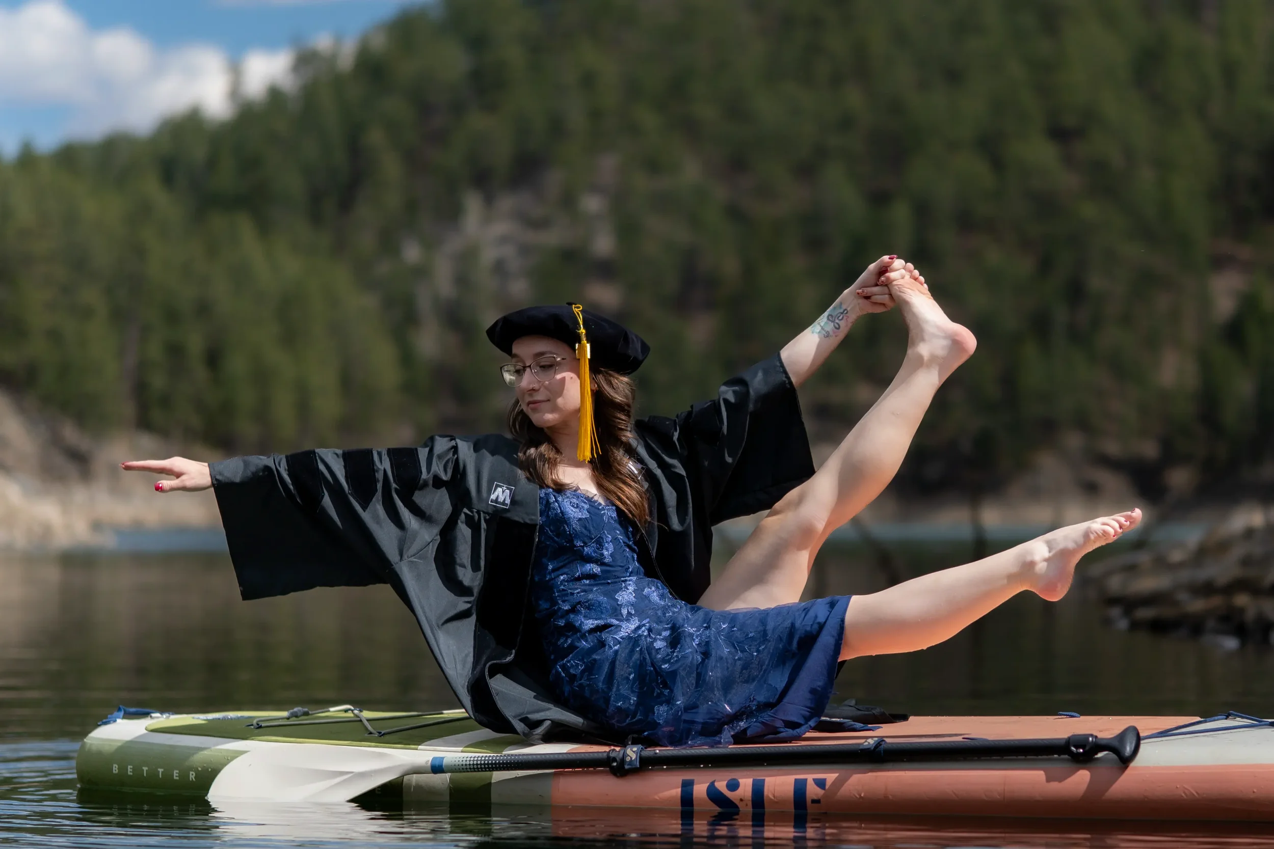 A woman in a graduation cap and gown doing a yoga pose on a paddleboard on a lake with a forested background.