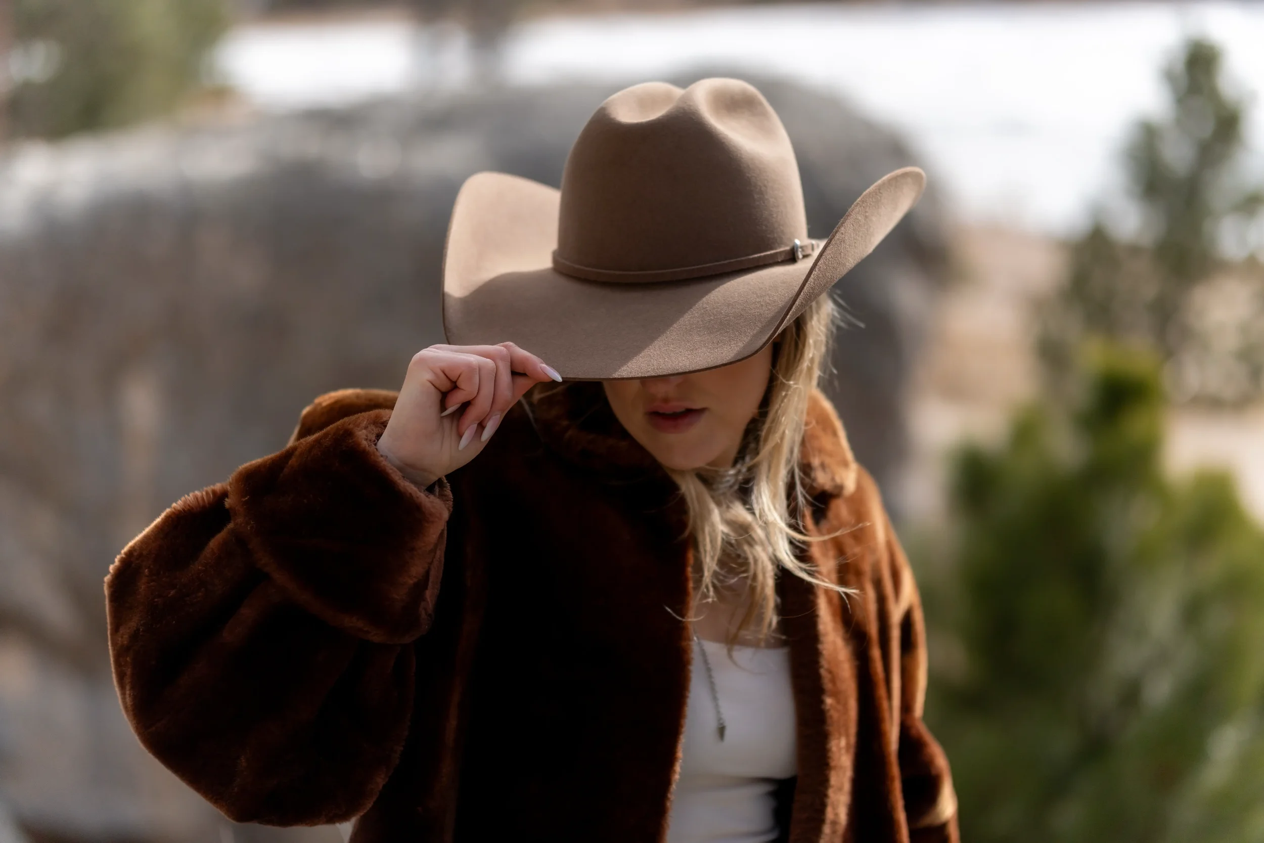 Woman wearing a large beige wide-brimmed hat and a brown fur coat outdoors in a natural setting.