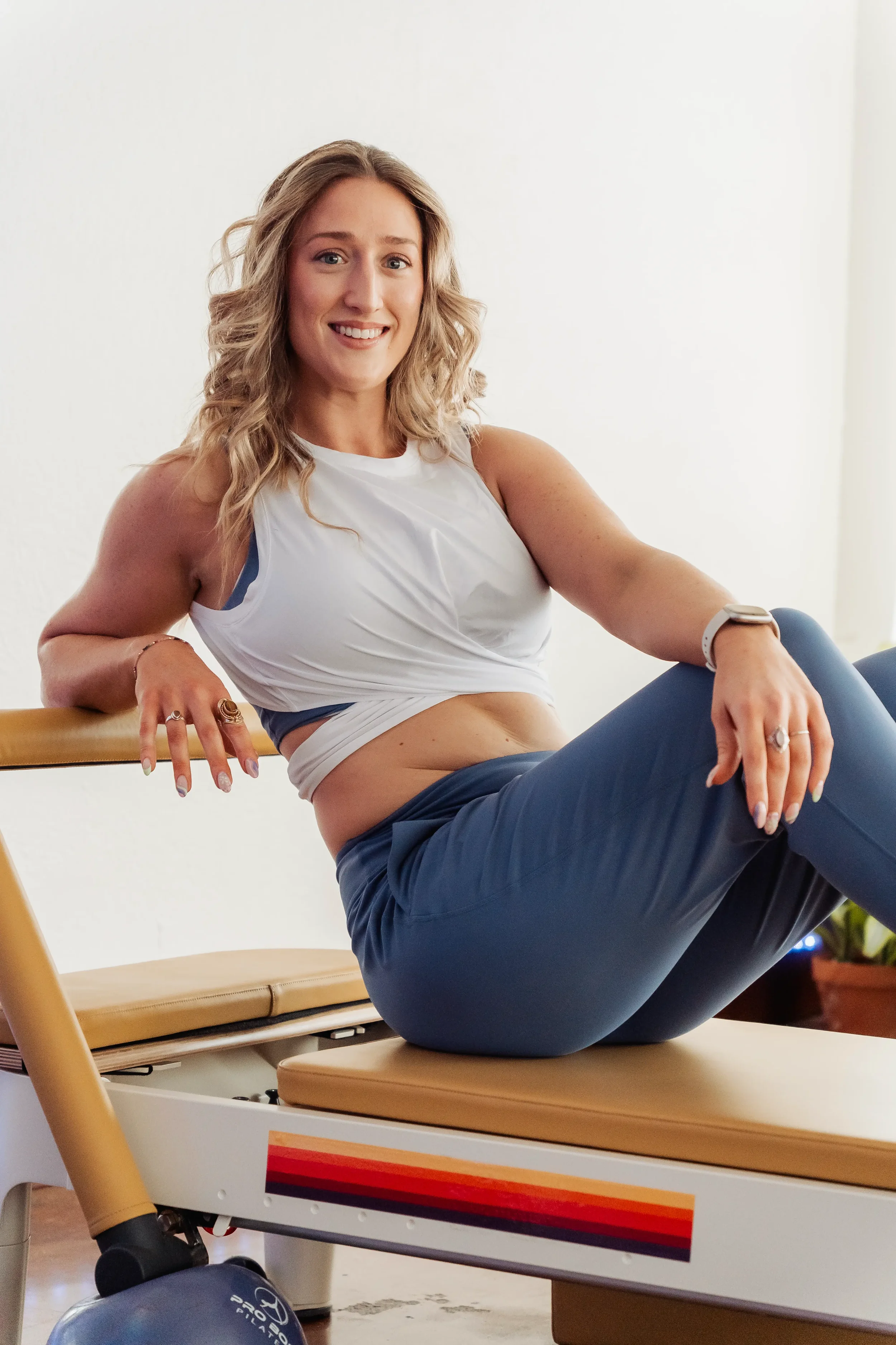 A woman with blonde, curly hair sitting on an exercise table, smiling, in a fitness or physical therapy setting, with a balance ball partially visible at the bottom of the image.