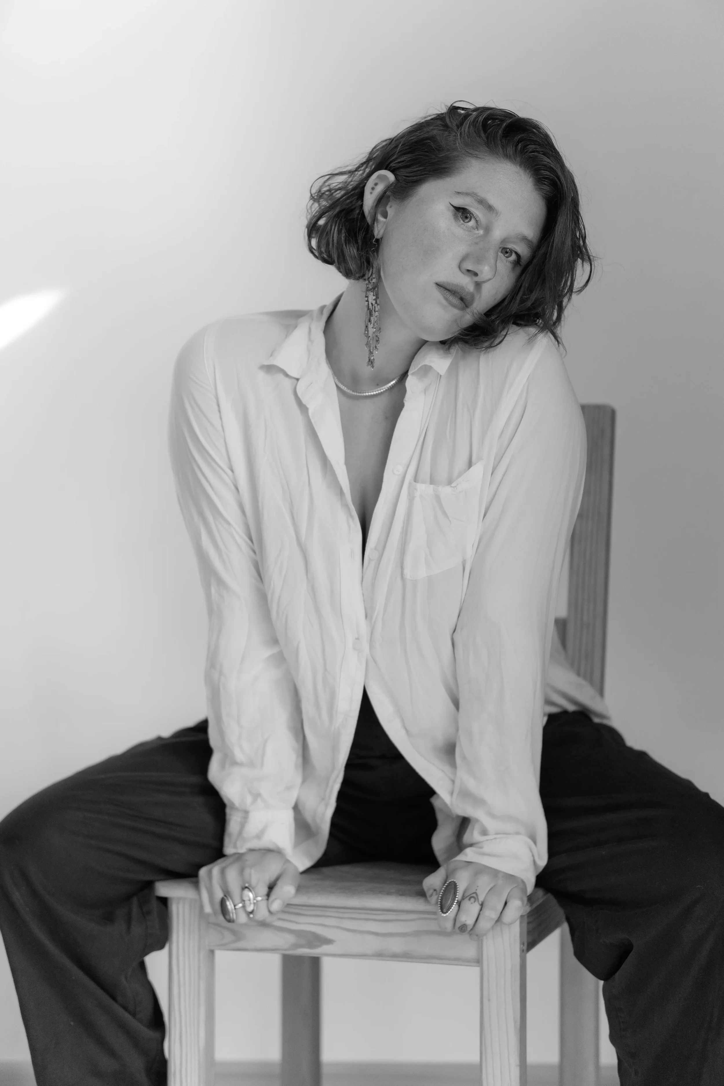Black and white photo of a young woman with short, wavy hair, sitting backwards on a wooden chair and leaning forward slightly, wearing a white shirt and dark pants, with her hands resting on the chair's back, looking at the camera.