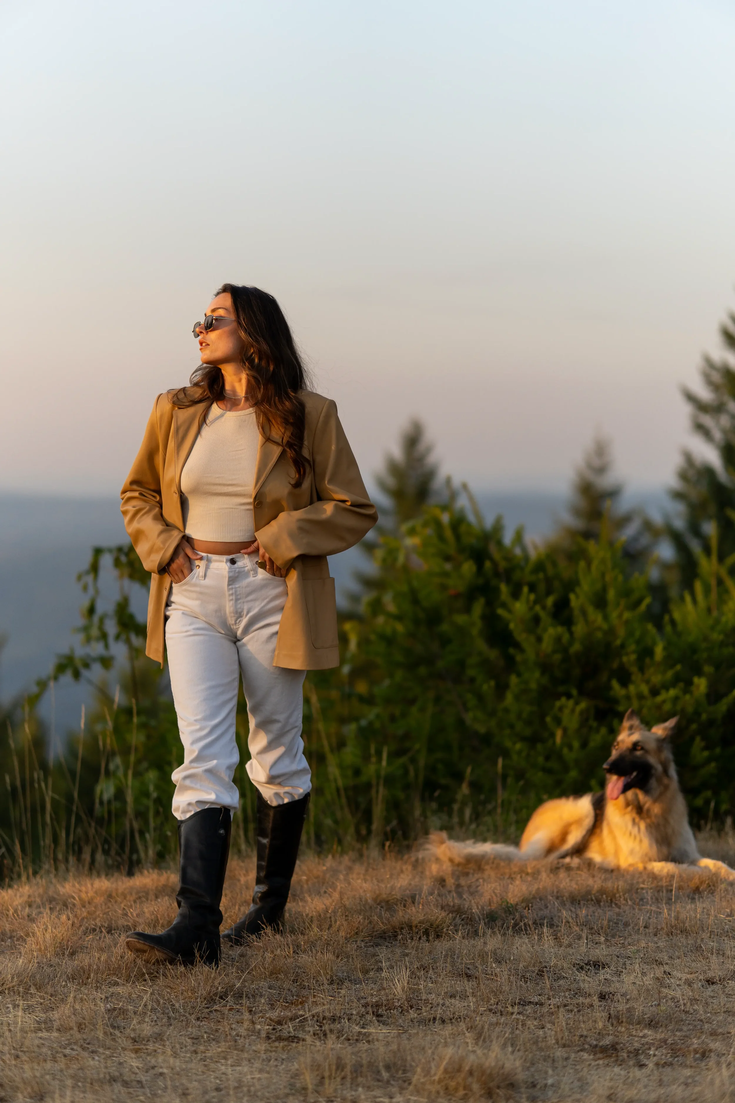 A woman with long wavy hair wearing sunglasses, a beige blazer, a white crop top, white pants, and black riding boots standing outdoors in a grassy area during sunset, with a German Shepherd dog lying on the ground nearby and trees in the background.
