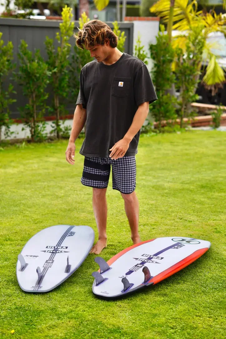 Young man with brown hair stands on grass in a backyard, looking at two surfboards lying on the ground.