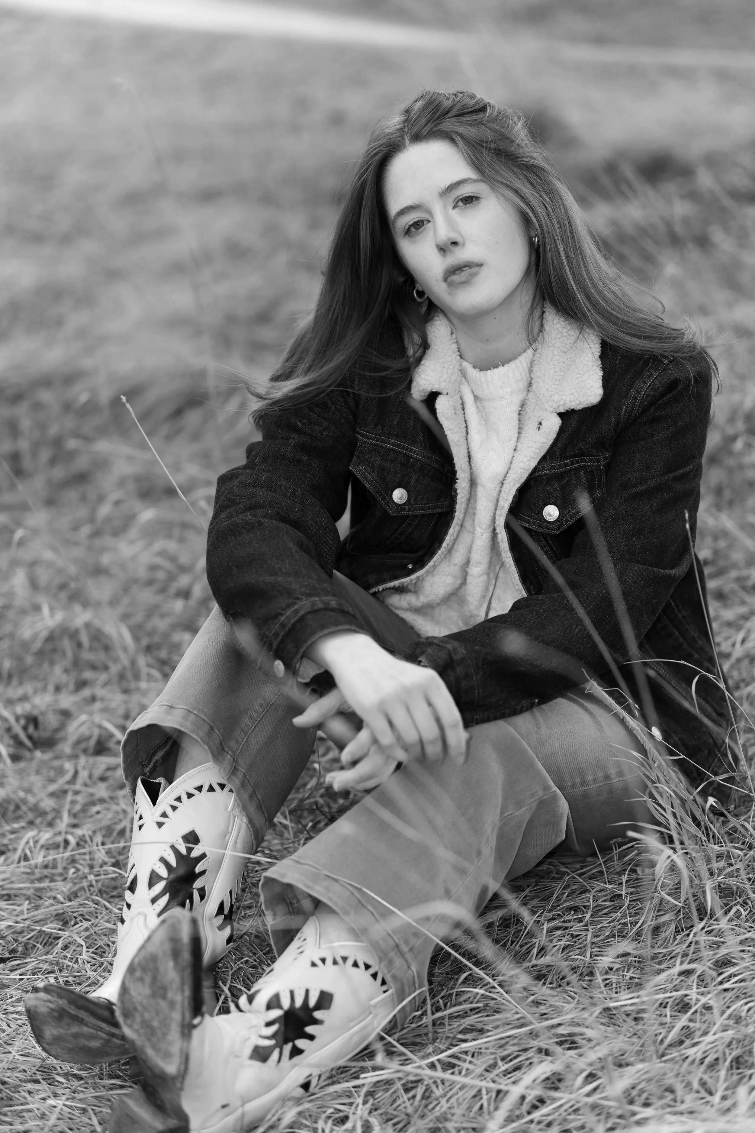 A young woman with long hair, dressed in a jacket and jeans, sitting on the grass in an outdoor setting, looking at the camera.