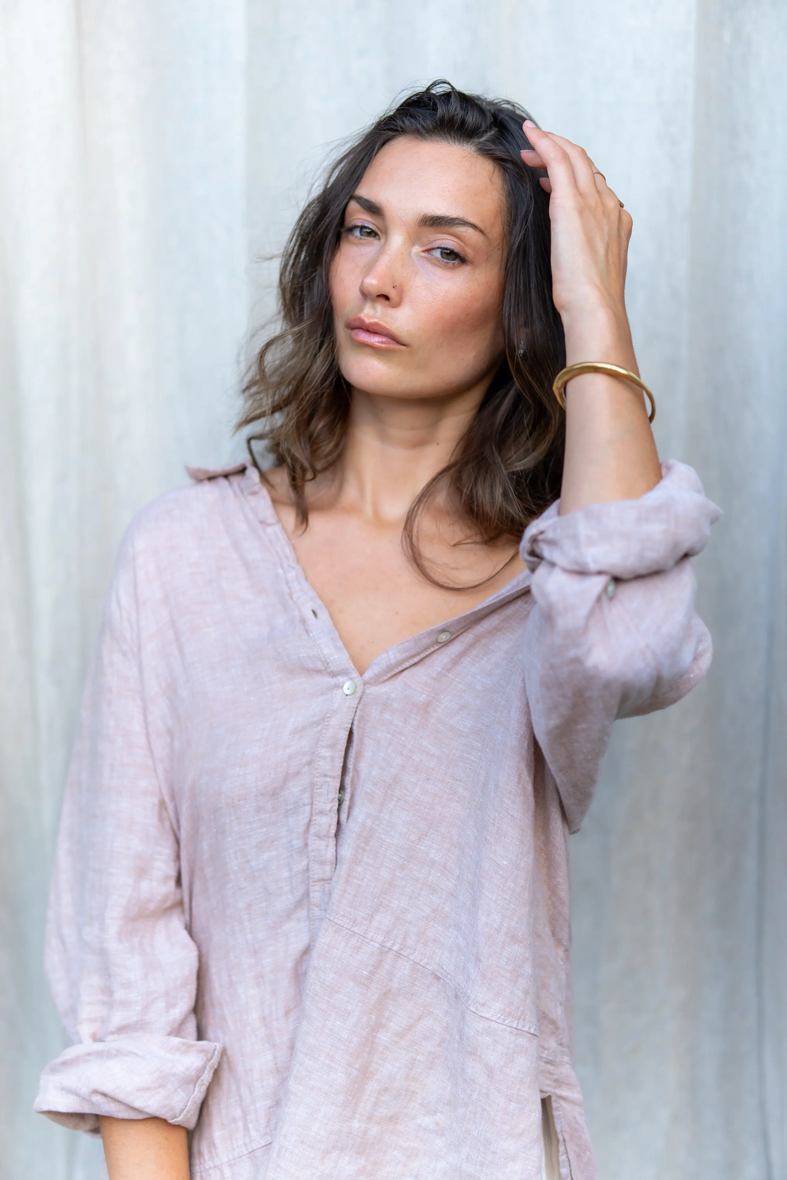 A woman with brown, wavy hair wearing a light pink button-down shirt with rolled-up sleeves, holding her hair back with one hand, and wearing a gold bracelet on her wrist, standing against a plain, light-colored background.