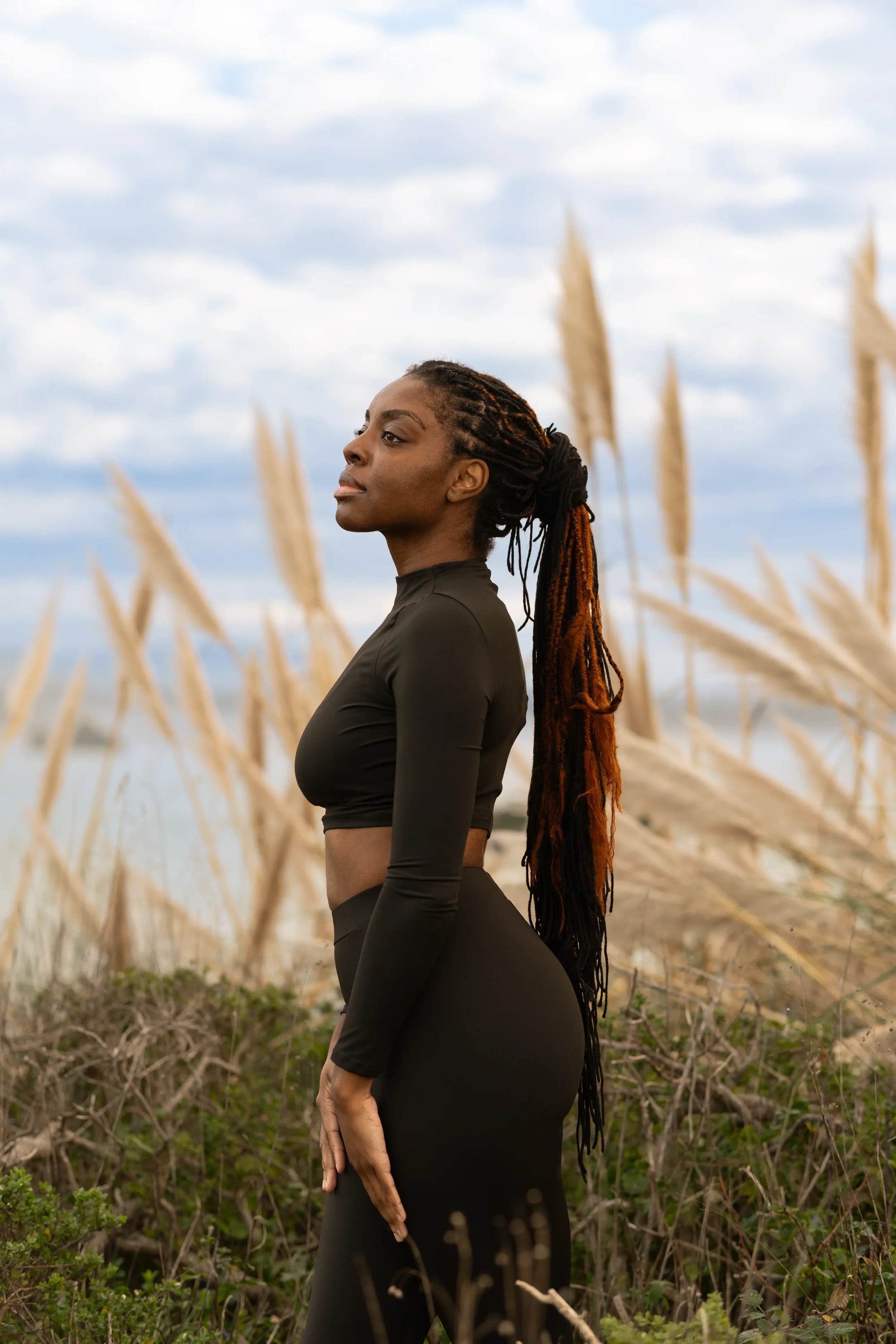 A woman with long braided hair wearing a black long-sleeve athletic outfit standing outdoors among tall brown grasses and greenery, with a partly cloudy sky in the background.