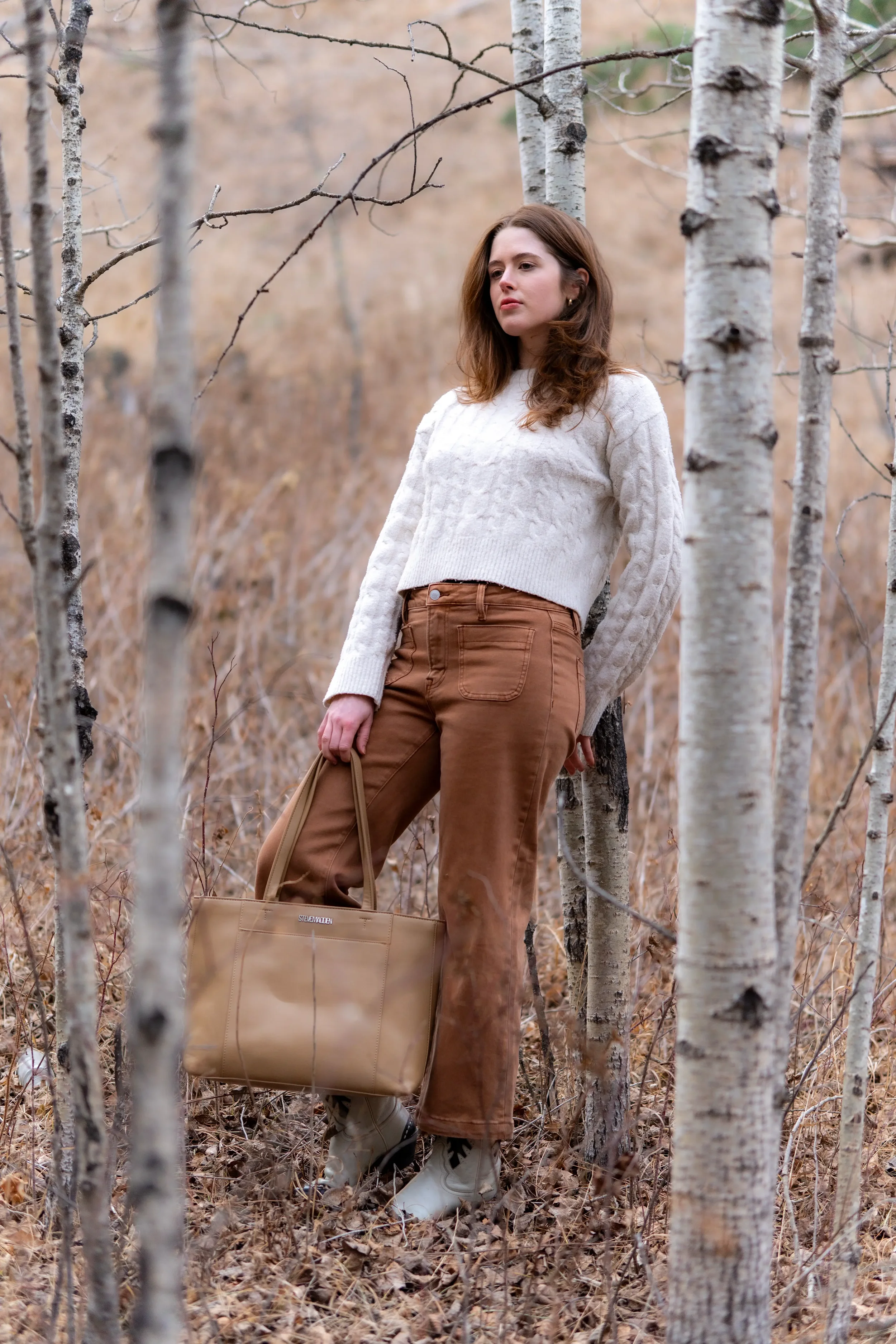 A woman with brown hair wearing a cream cable knit sweater and brown pants stands among leafless trees in a forest, holding a tan tote bag and wearing beige and black boots.