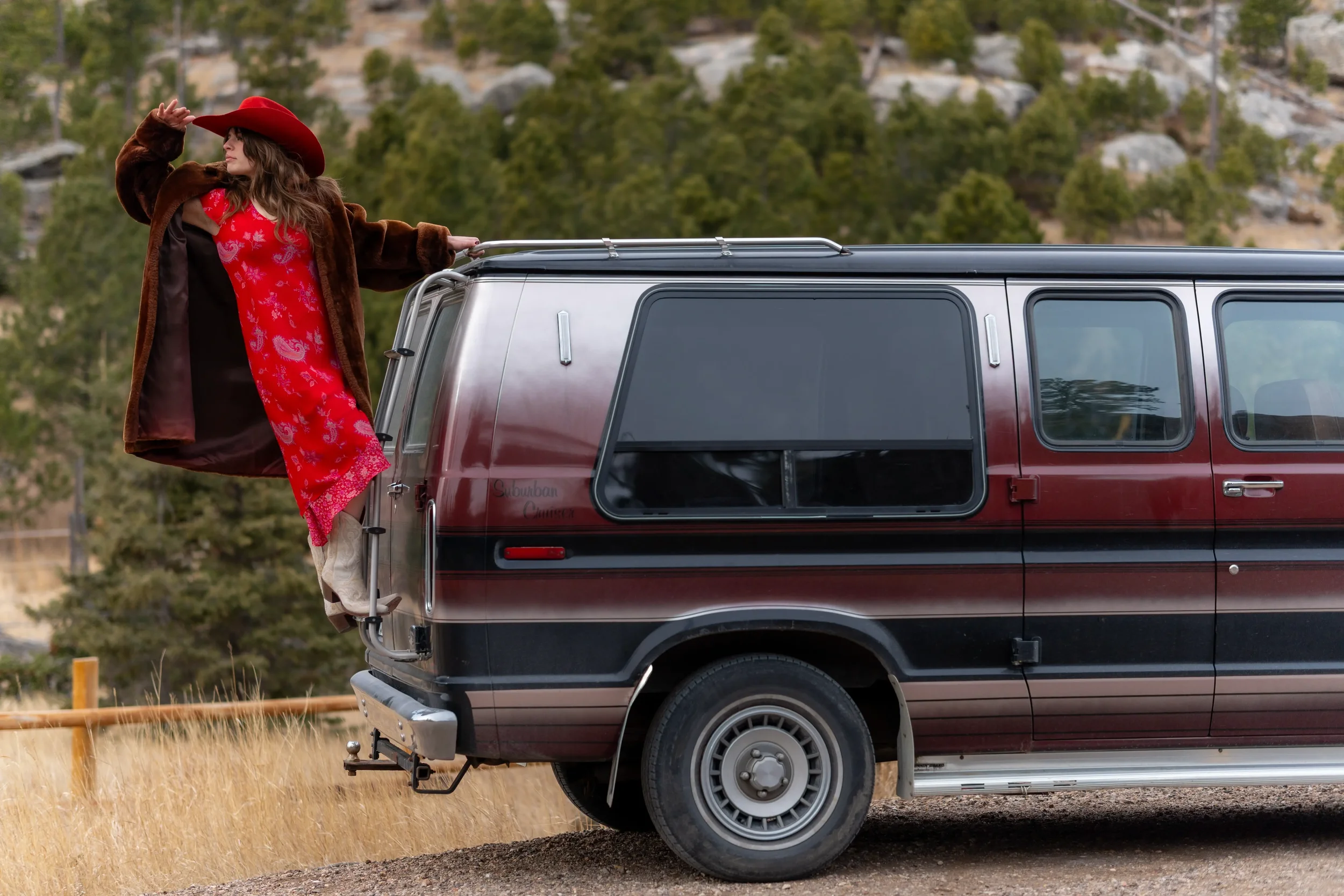 Woman in a red dress, brown coat, and red hat standing on the side of a red camper van in a grassy outdoor area with trees in the background.