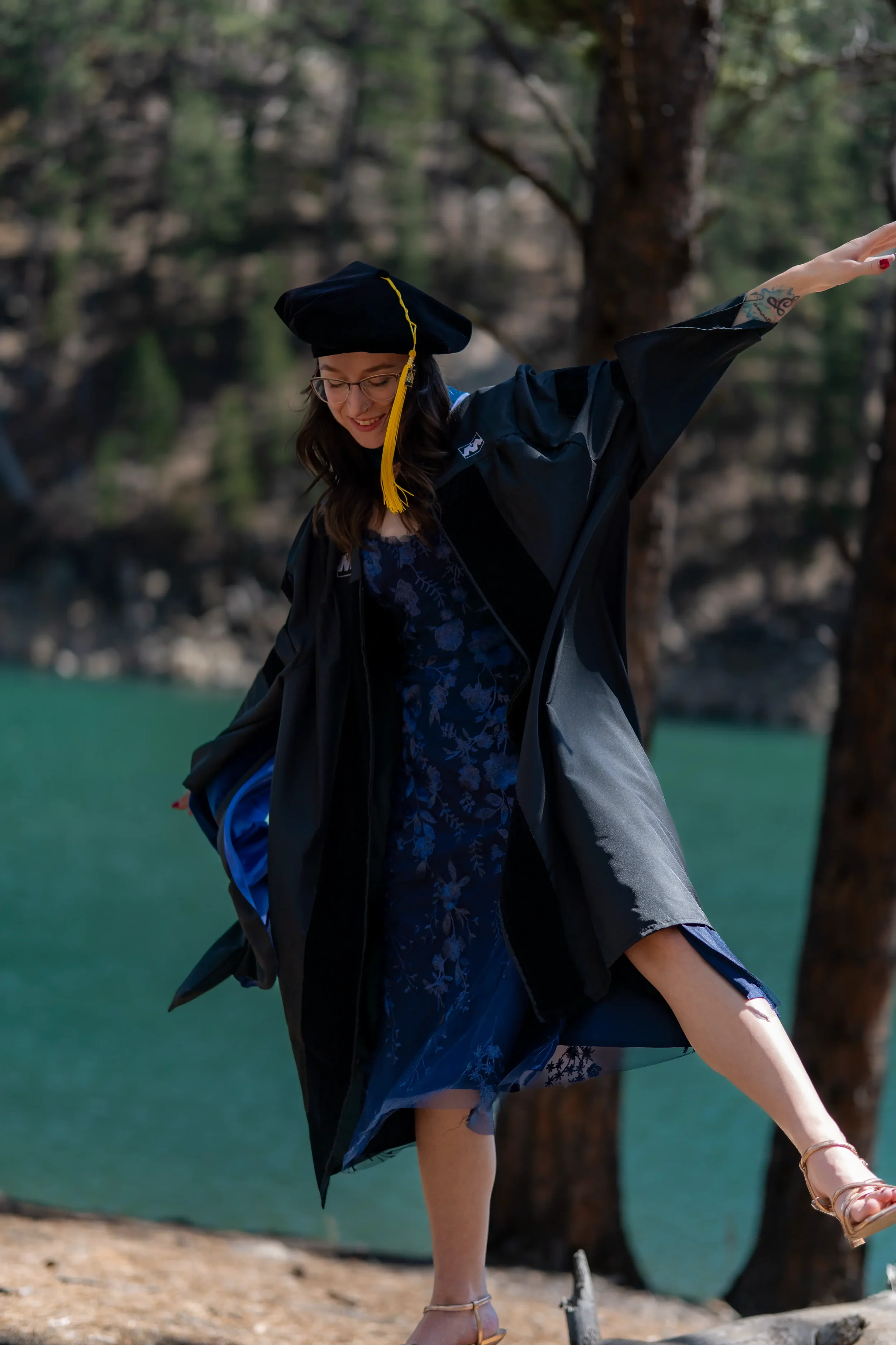 A woman in a graduation cap and gown standing outdoors near a lake, smiling, with her arm extended outward and her leg raised.