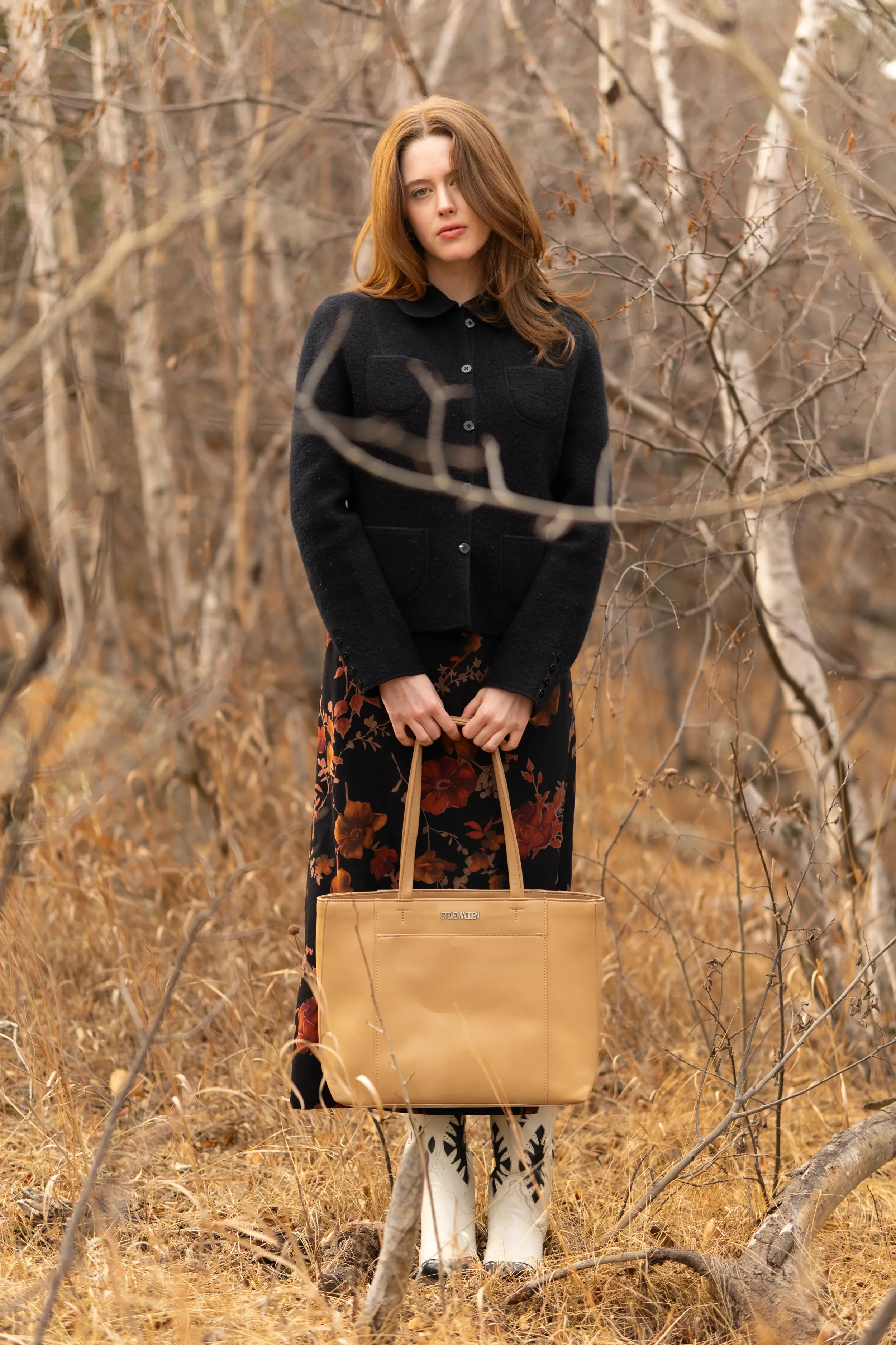 A woman with long red hair wearing a black jacket stands in a dry, leafless forest holding a tan tote bag. She is wearing white cowboy boots with black designs and a floral skirt.