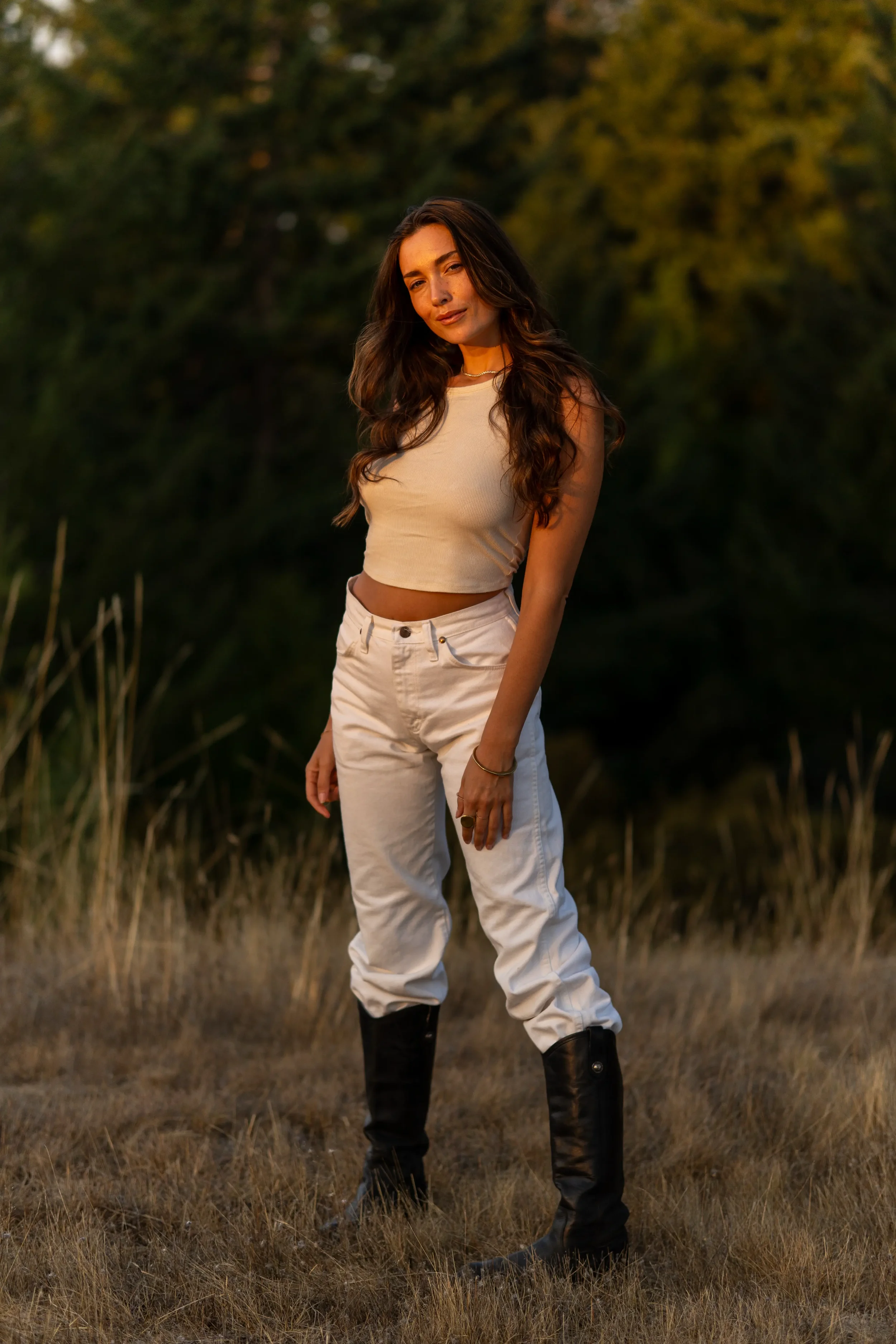 A woman standing outdoors in a grassy area with trees in the background during golden hour, wearing a beige crop top, white pants, and black boots.
