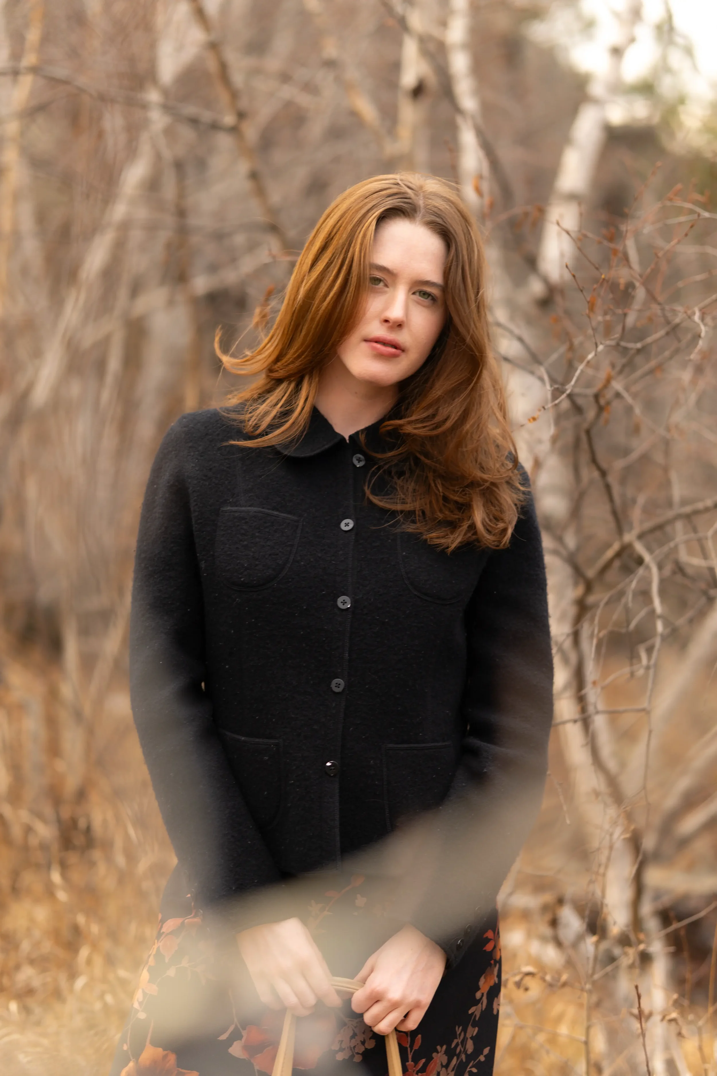A woman with red hair wearing a black coat stands outdoors among leafless trees, holding a paper bag.