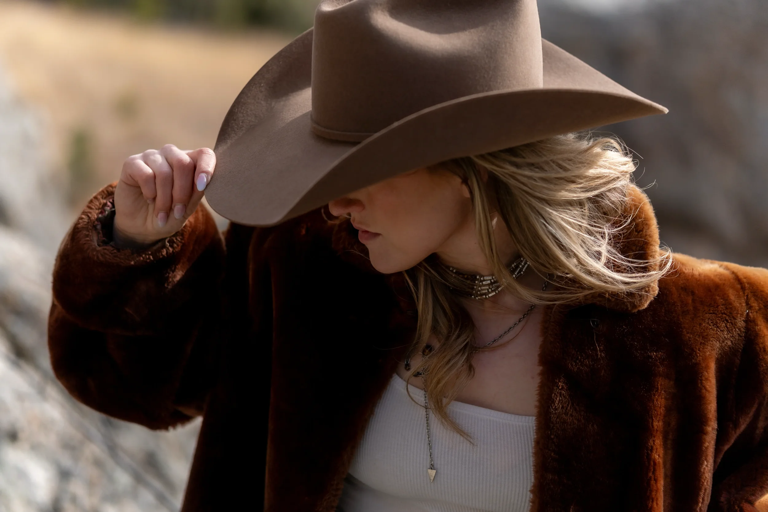 A woman wearing a large tan cowboy hat, brown fur coat, white top, and layered necklaces. She is holding her hat with her right hand and looking down.