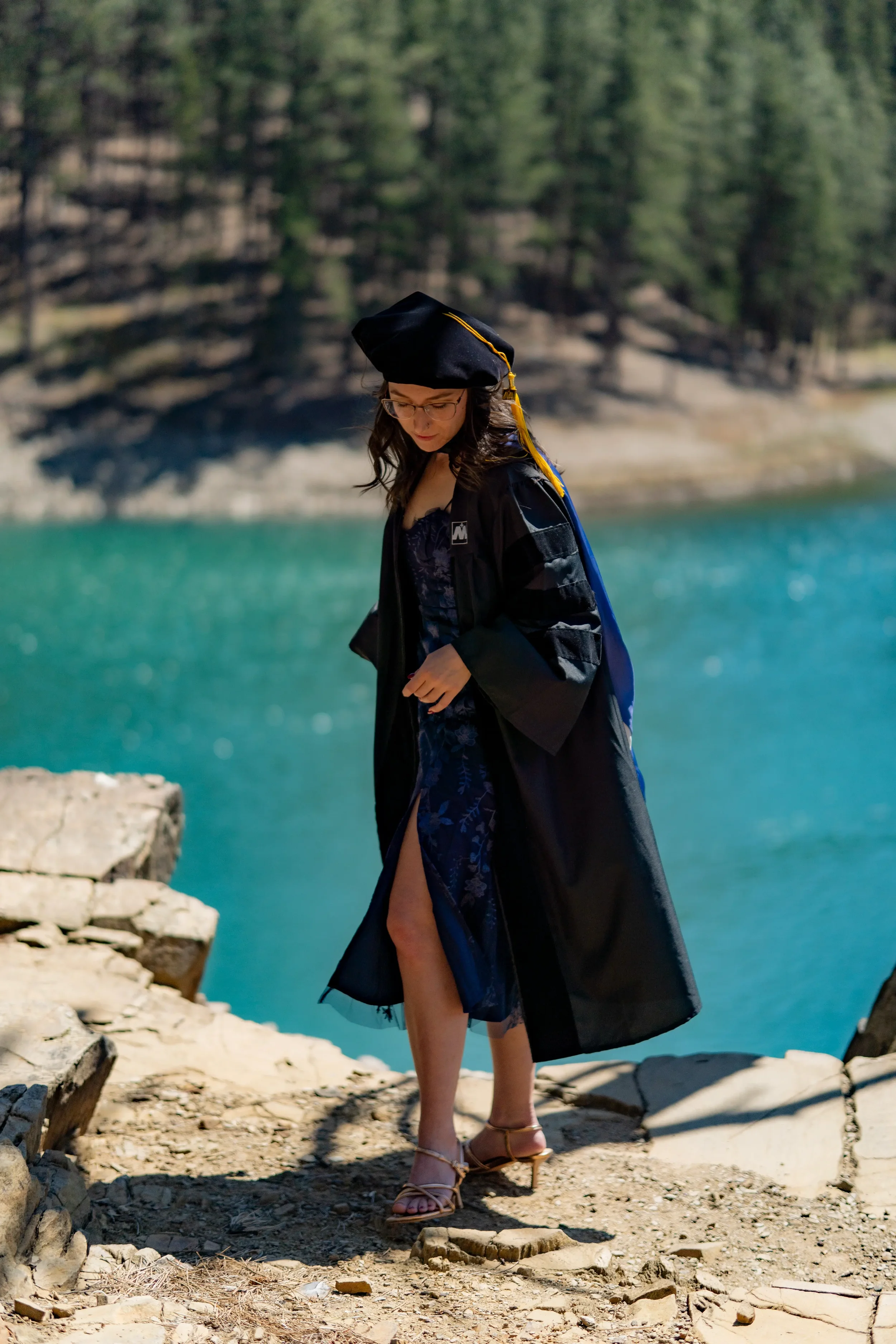 Woman in graduation gown and cap standing on rocky shoreline near turquoise water with trees in background
