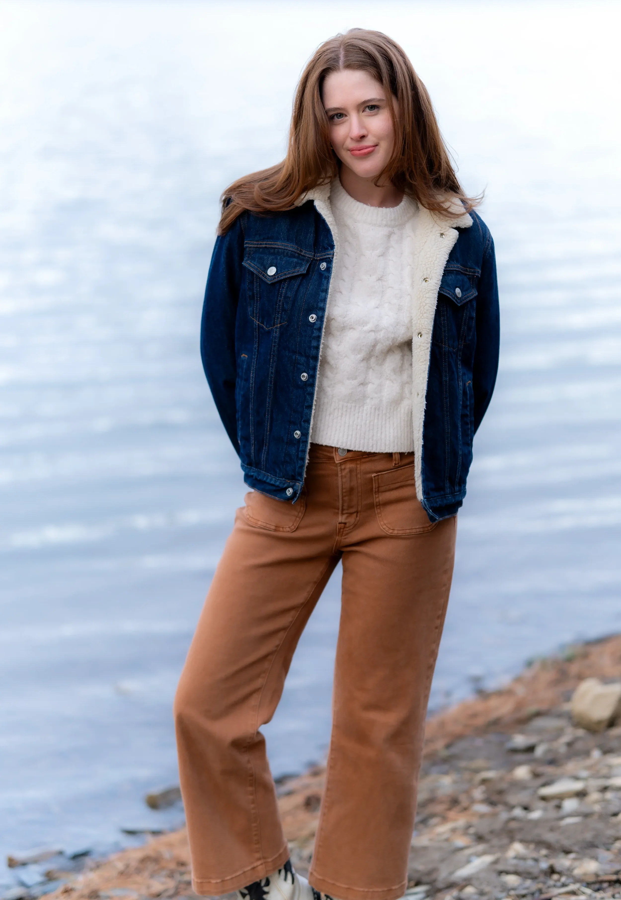 A young woman with long brown hair standing outdoors near a body of water, wearing a denim jacket with a shearling lining, a white sweater, and brown pants.