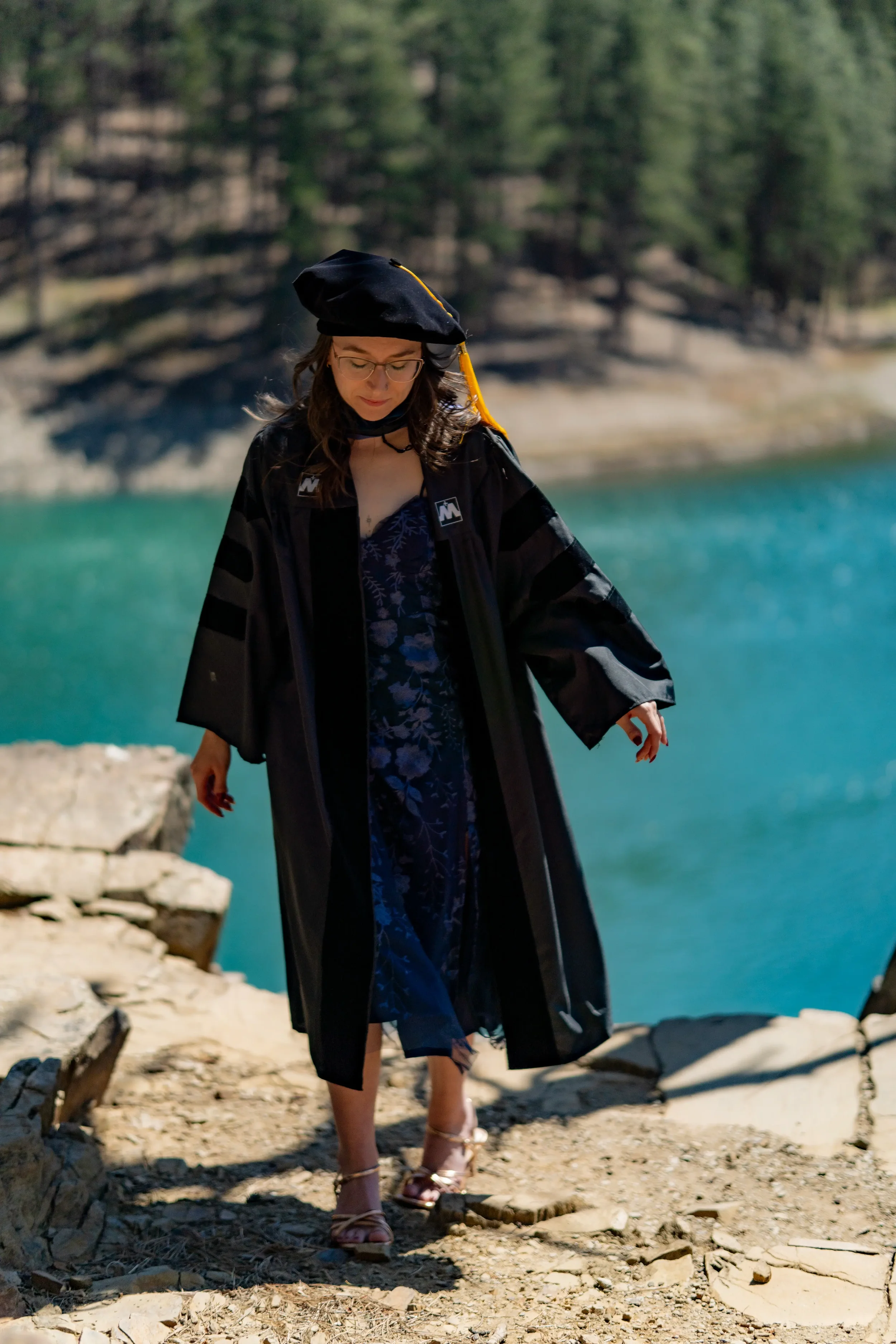 A woman wearing graduation attire walking on a rocky trail near a body of water and surrounded by trees.